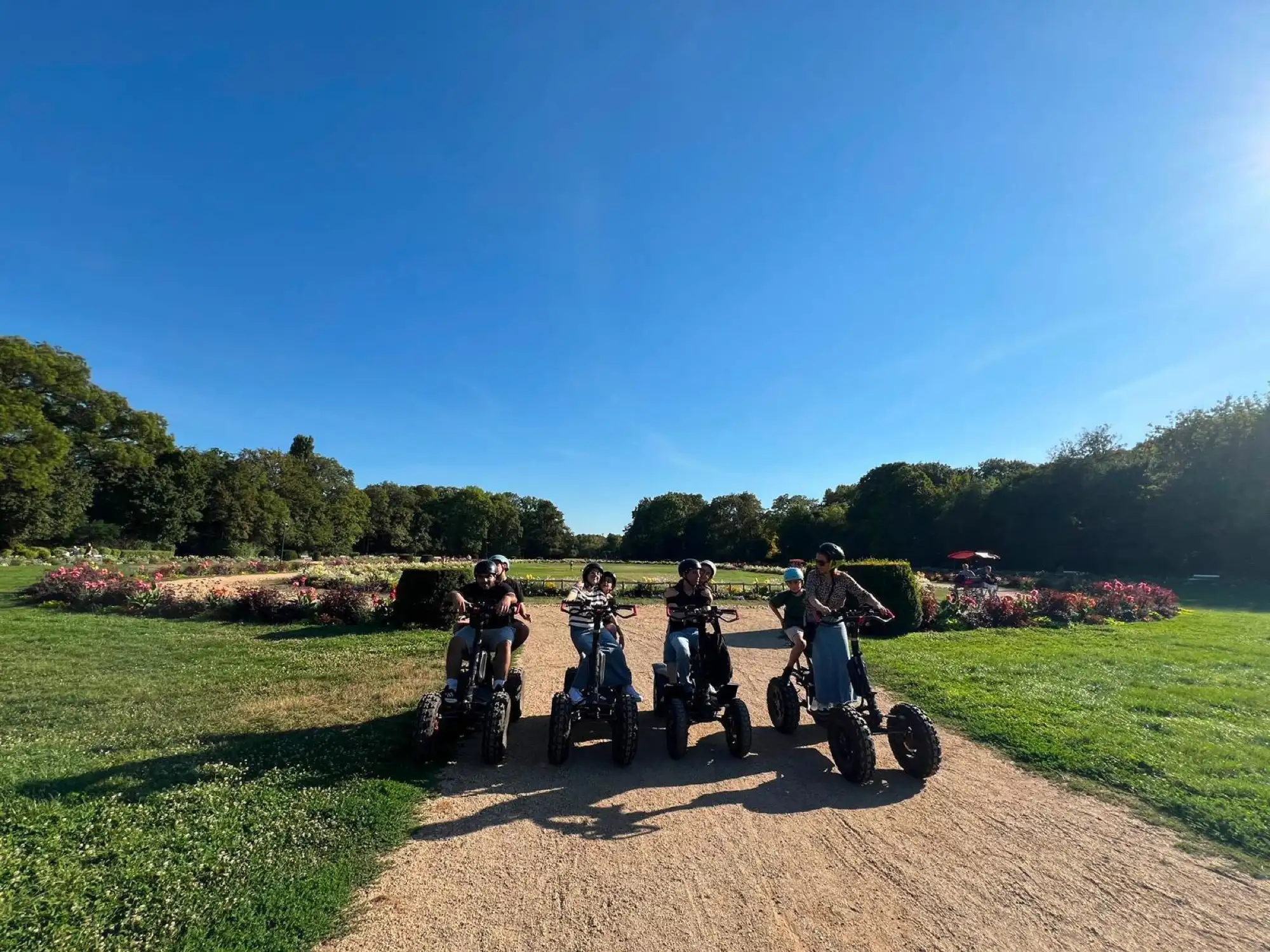Four riders on EZRaider electric four-wheelers lined up on a gravel path across a lawn on Margaret Island, with flower beds and trees behind