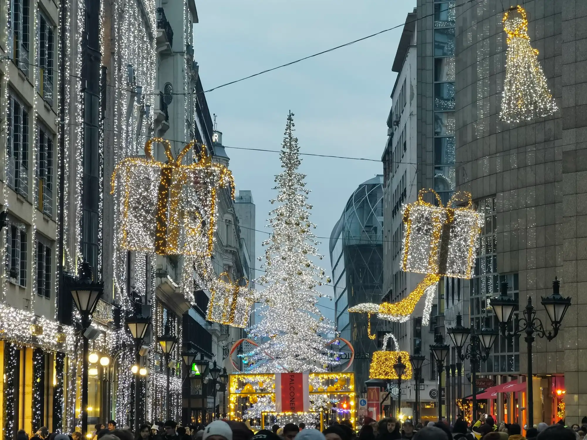 A giant Christmas tree made of white lights in the middle of Deák Ferenc utca, with huge lit gift boxes and a lit high-heeled shoe hanging over the street and light curtains on the buildings