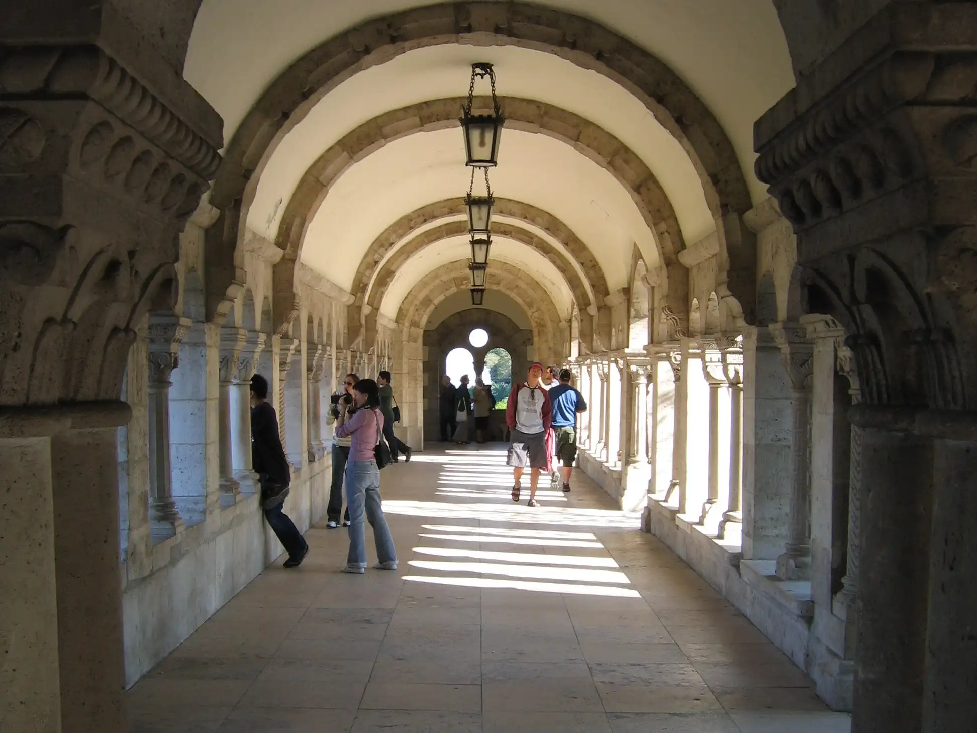 Visitors photographing the view through the columns of a vaulted stone arcade at Fisherman's Bastion, with lanterns hanging from the ceiling