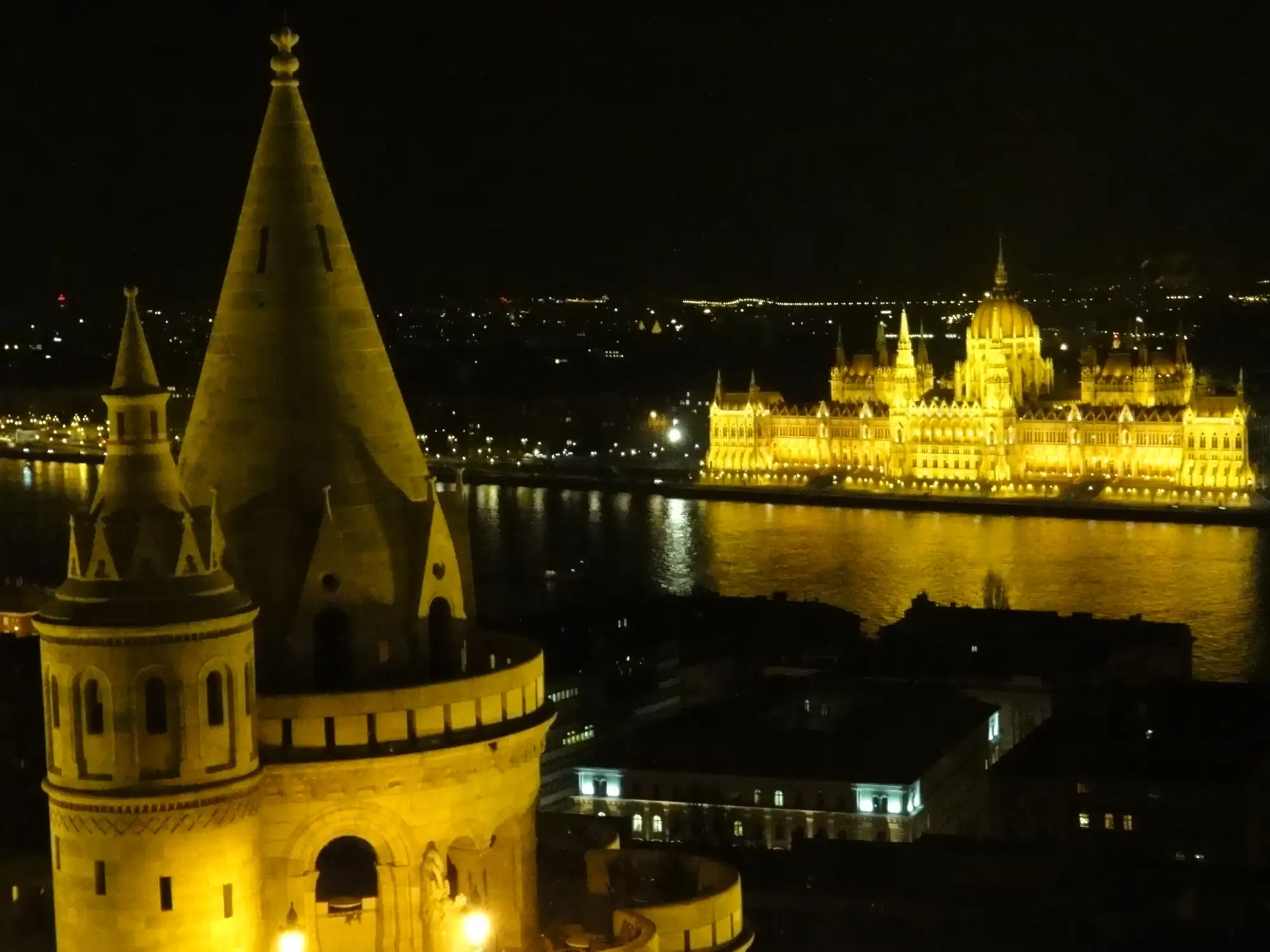 A floodlit conical tower of Fisherman's Bastion at night, with the illuminated Hungarian Parliament reflected in the Danube beyond