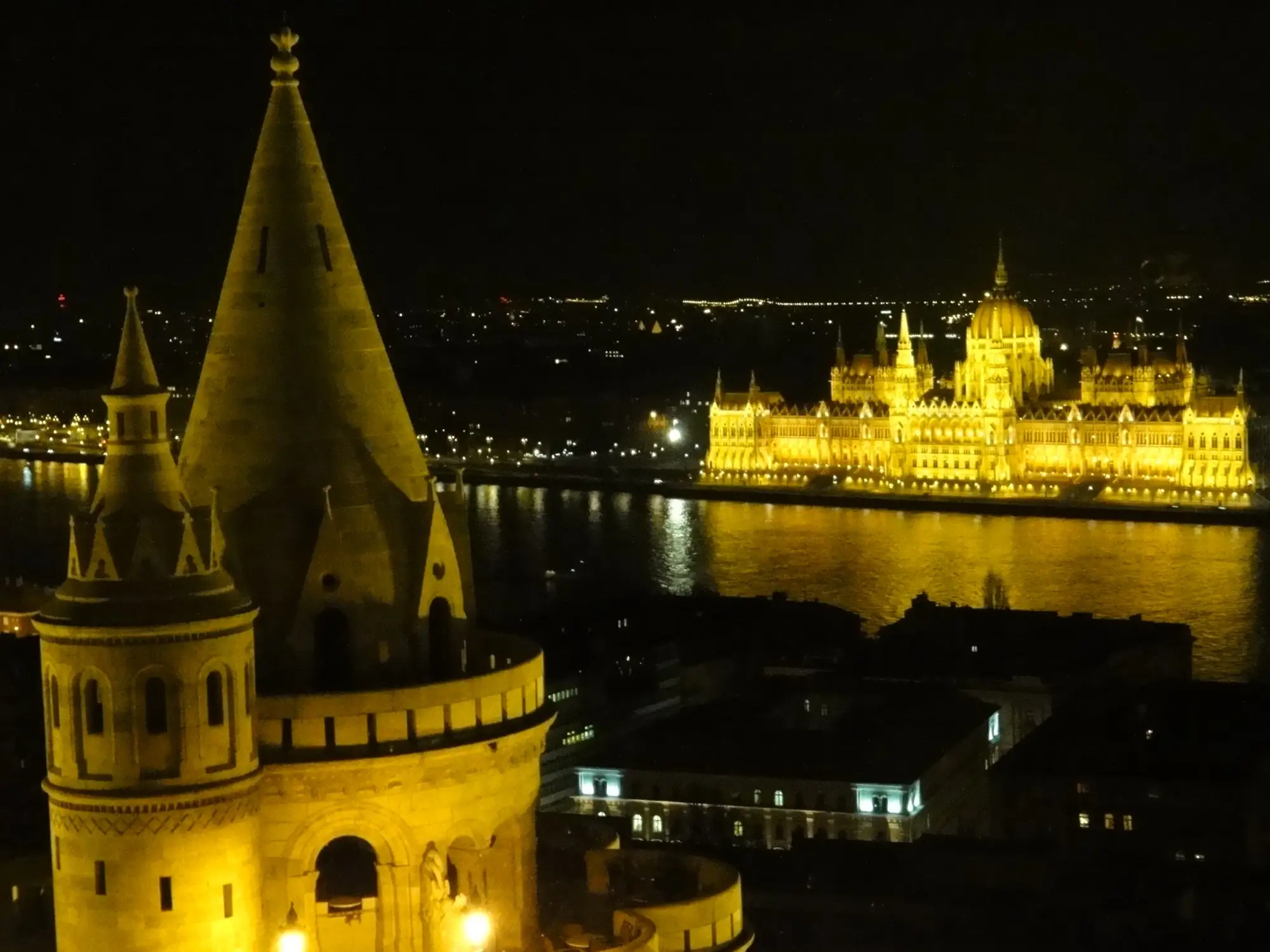 A floodlit conical tower of Fisherman's Bastion at night, with the illuminated Hungarian Parliament reflected in the Danube beyond