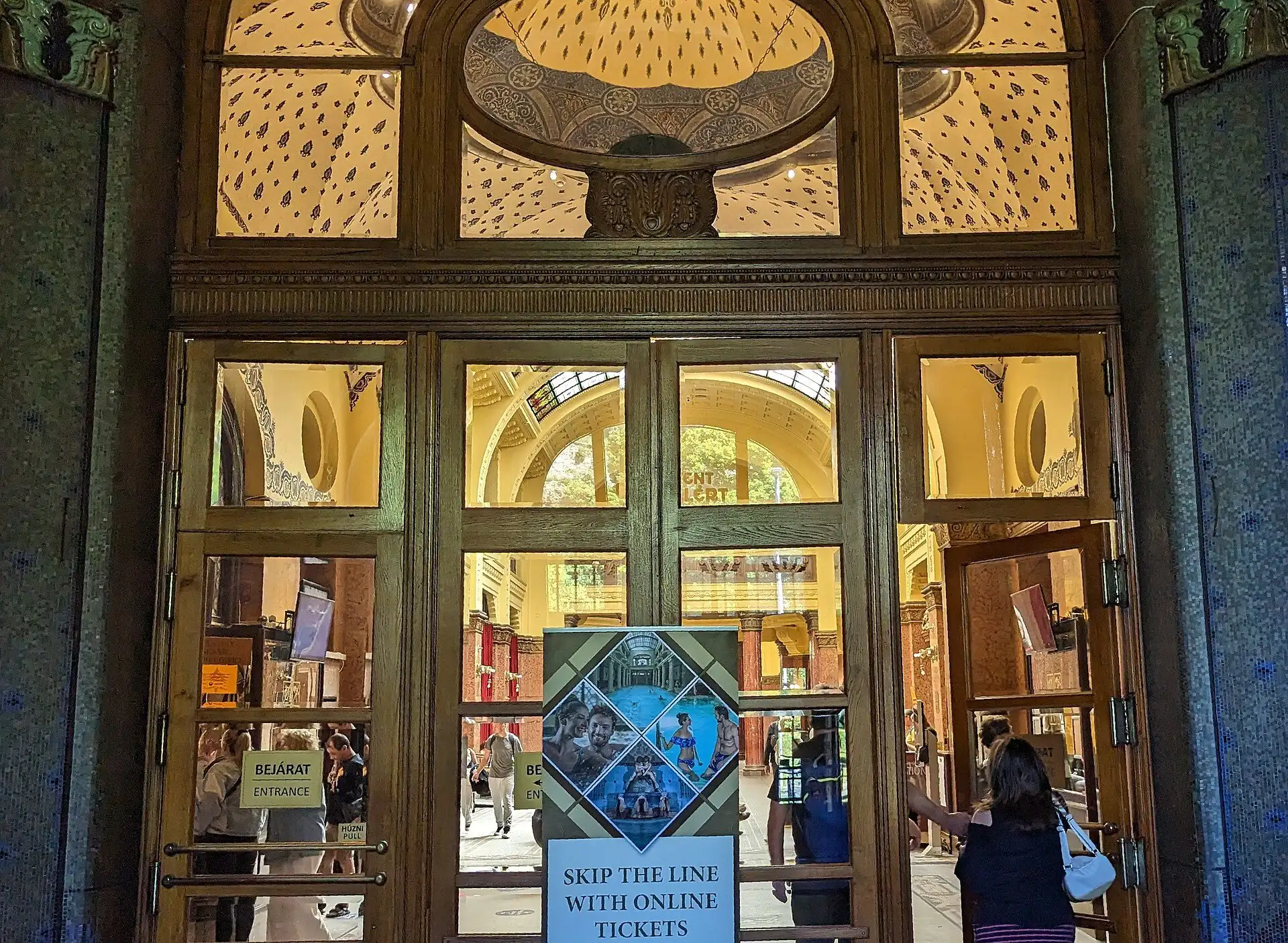 The entrance doors of Gellért Thermal Bath, dark wood and glass under a carved stone arch with stained glass above