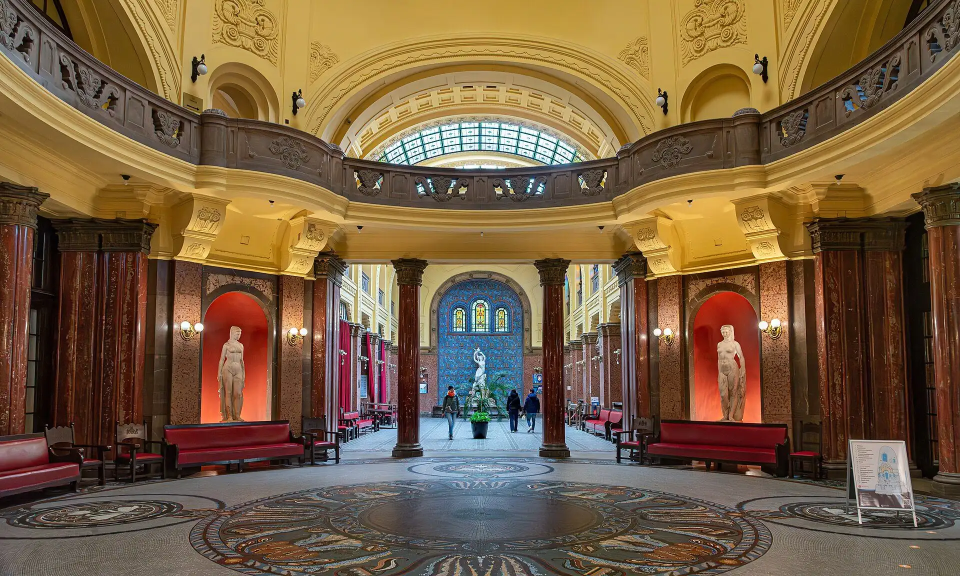 The entrance hall of Gellért Thermal Bath, with its dome, stone gallery, red marble columns and statues in orange-lit niches