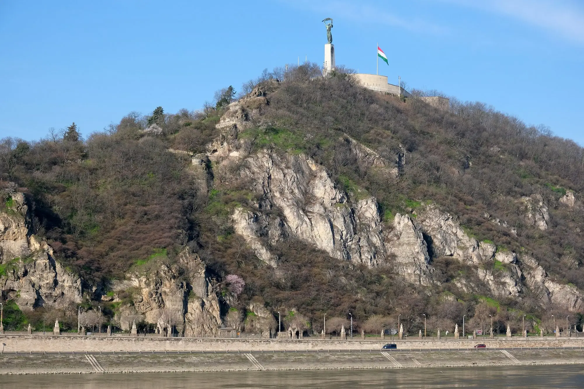 The rocky south face of Gellért Hill rising above the Danube embankment in Budapest, with the Liberty Statue and the national flag on the summit