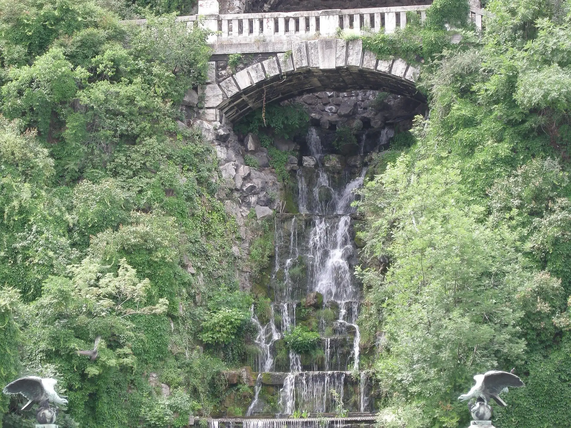 The artificial waterfall below the St Gellért monument running, water cascading down the rock from under a stone arch between thick greenery, with the bronze eagles on their columns at the bottom