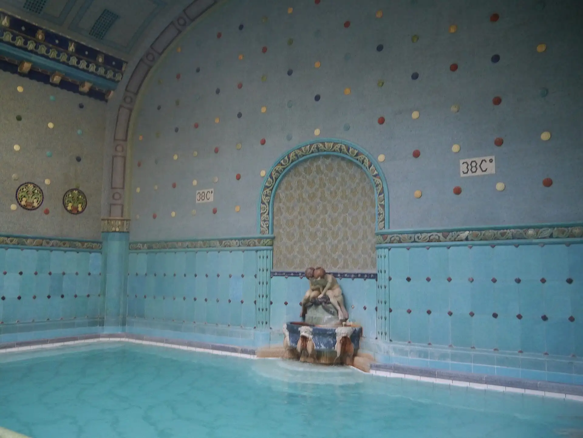 A thermal pool in the blue Zsolnay-tiled hall at Gellért Thermal Bath, with a fountain in an arched niche and a 38°C sign on the wall