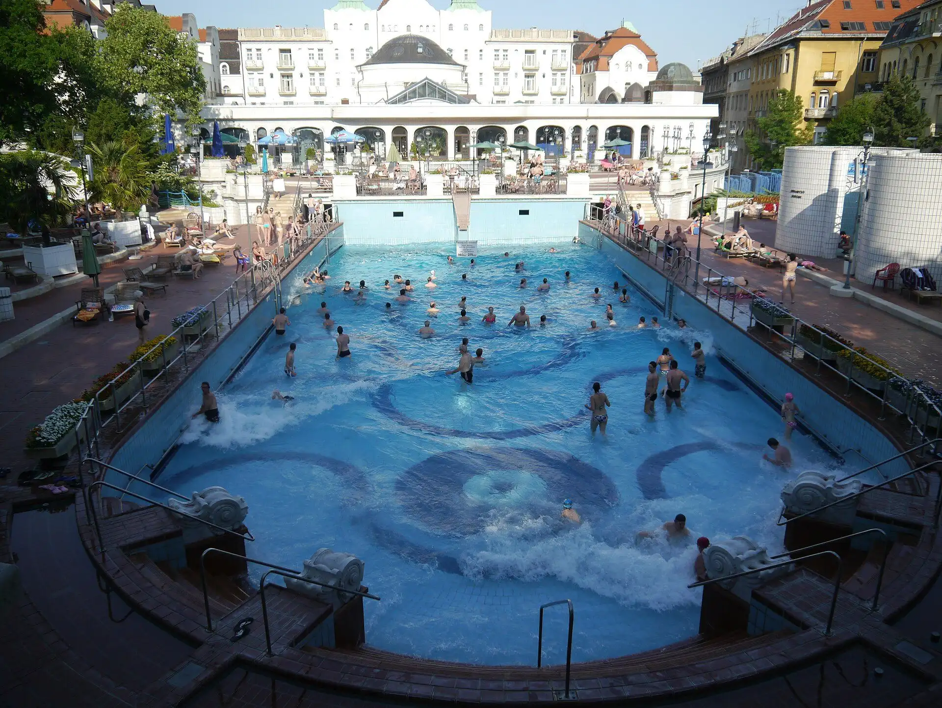 The outdoor wave pool at Gellért Thermal Bath with waves running and bathers in the water, the hotel and its domes behind