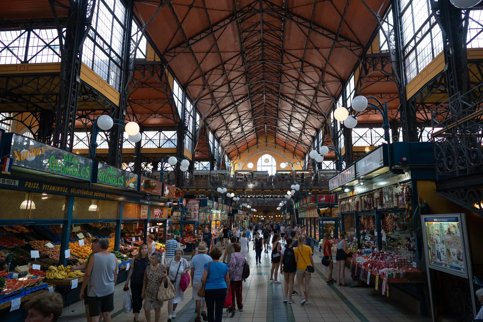 The main aisle on the ground floor of the Great Market Hall in Budapest, with produce stalls on the left and shoppers walking down the nave