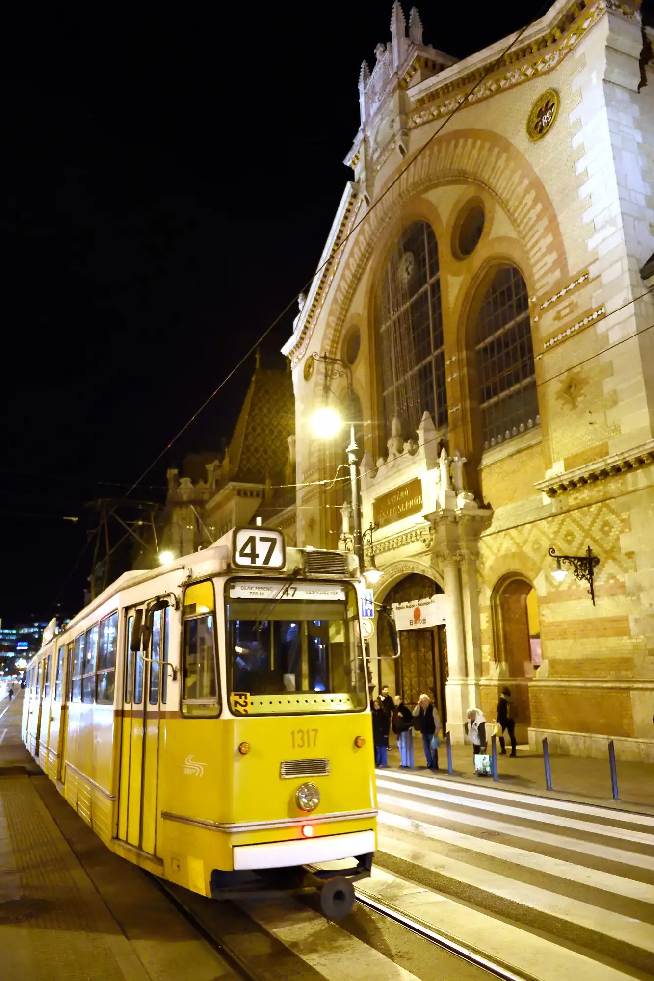 A yellow tram on route 47 stopping outside the lit front of the Great Market Hall in Budapest at night