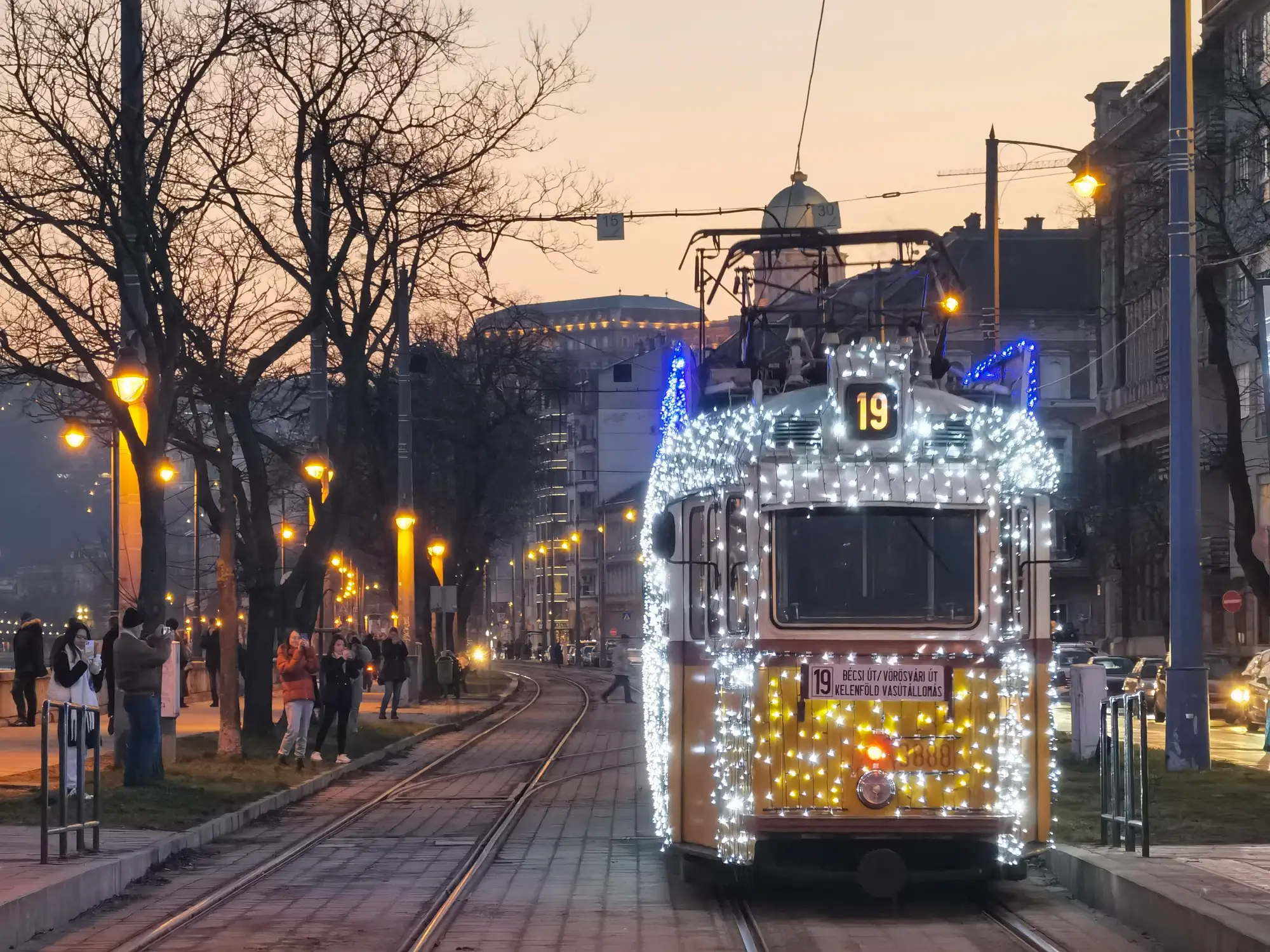 An old Budapest tram covered in white Christmas lights on line 19 at Batthyány tér at dusk, with the Danube promenade lamps lit beside it