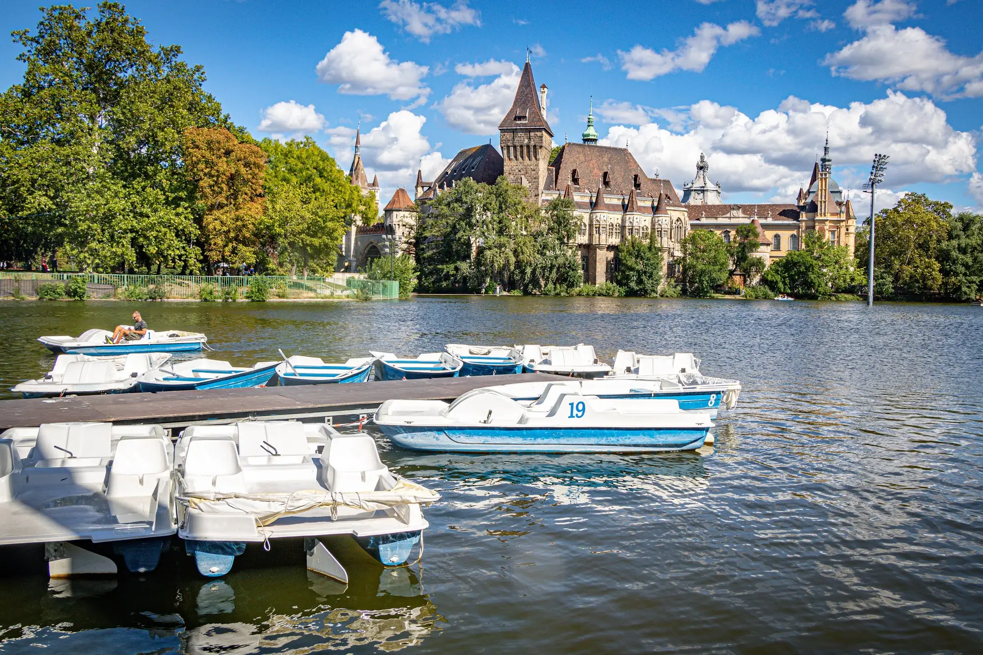 Pedal boats moored at a jetty on the City Park boating lake in Budapest in summer, with Vajdahunyad Castle rising above the trees on the far bank