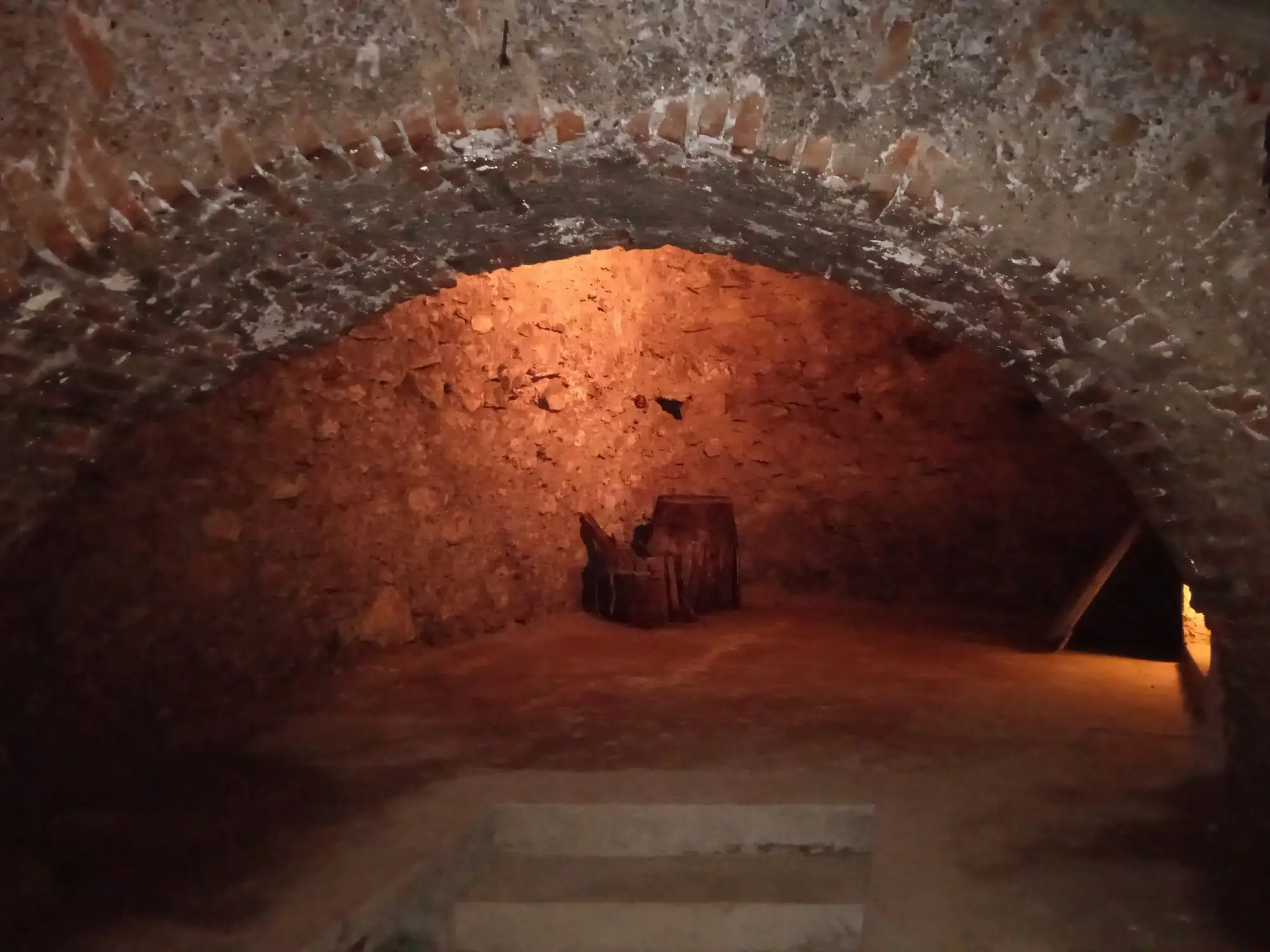 A brick and stone barrel-vaulted cellar in the Buda Castle Cave, lit warm, with a wooden barrel against the far wall