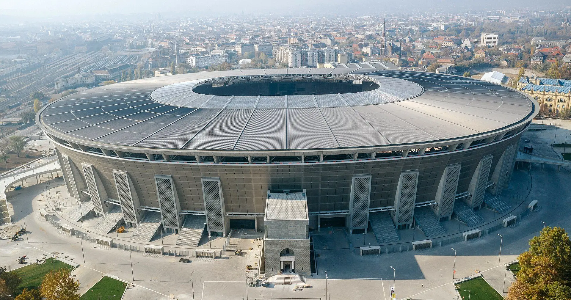 Puskás Aréna from the air, the round grey stadium with its full roof, the Puskás Aréna Park around it and Budapest beyond
