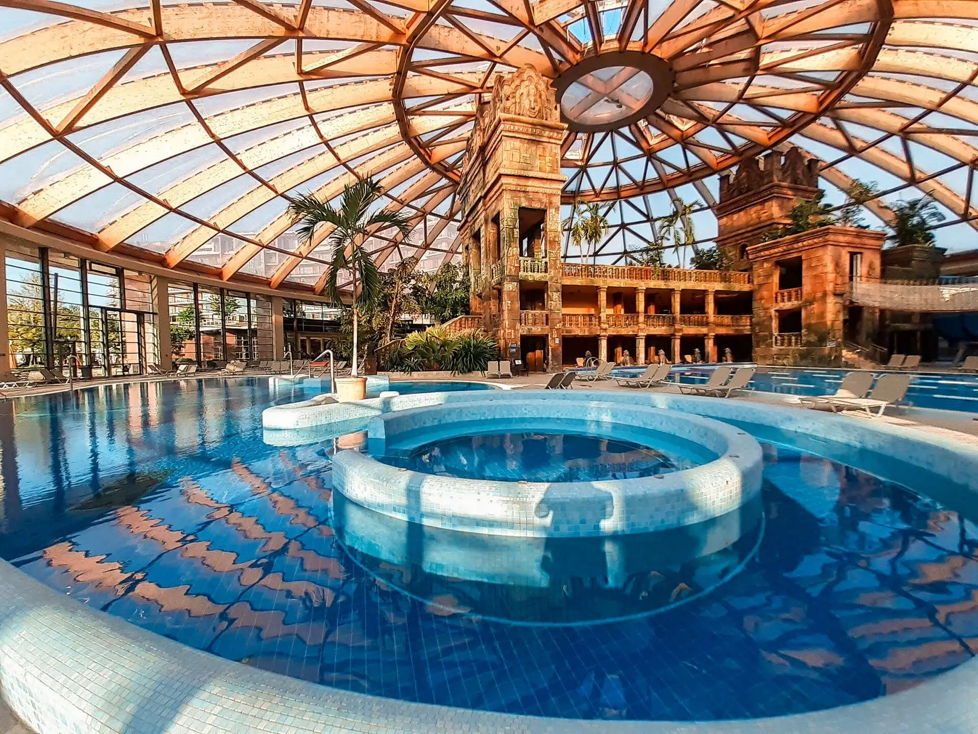 The adventure pool and a round jacuzzi at Aquaworld, under the see-through dome and its timber ribs, with the Angkor-style temple behind