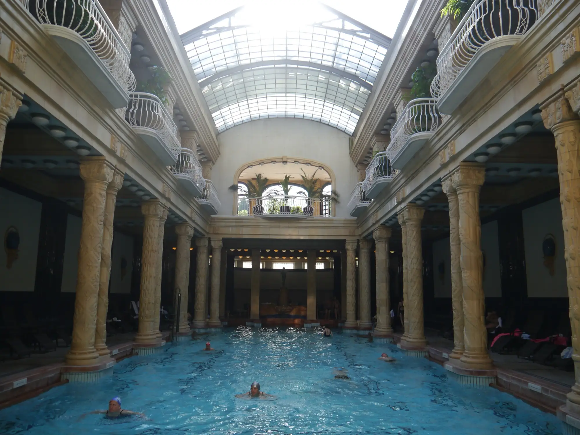 The indoor swimming pool hall of Gellért Thermal Bath in Budapest, with carved columns, balconies with white railings and a glass roof over the pool
