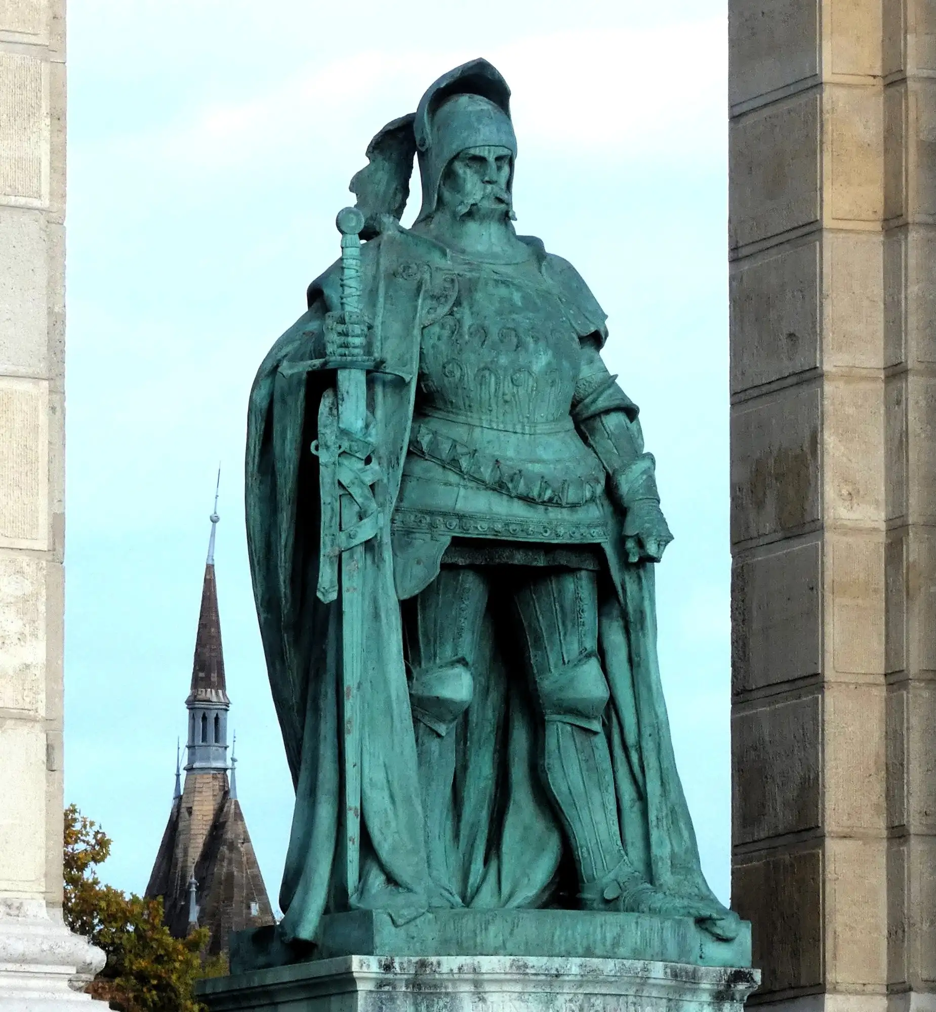 A bronze statue of a Hungarian ruler in armour, holding a sword, standing between the stone columns of the Heroes' Square colonnade