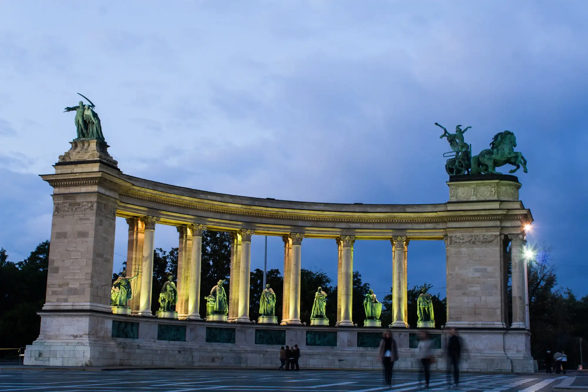 The left-hand colonnade of the Millennium Monument lit at dusk, seven bronze kings standing between its columns and the War chariot on its right-hand end