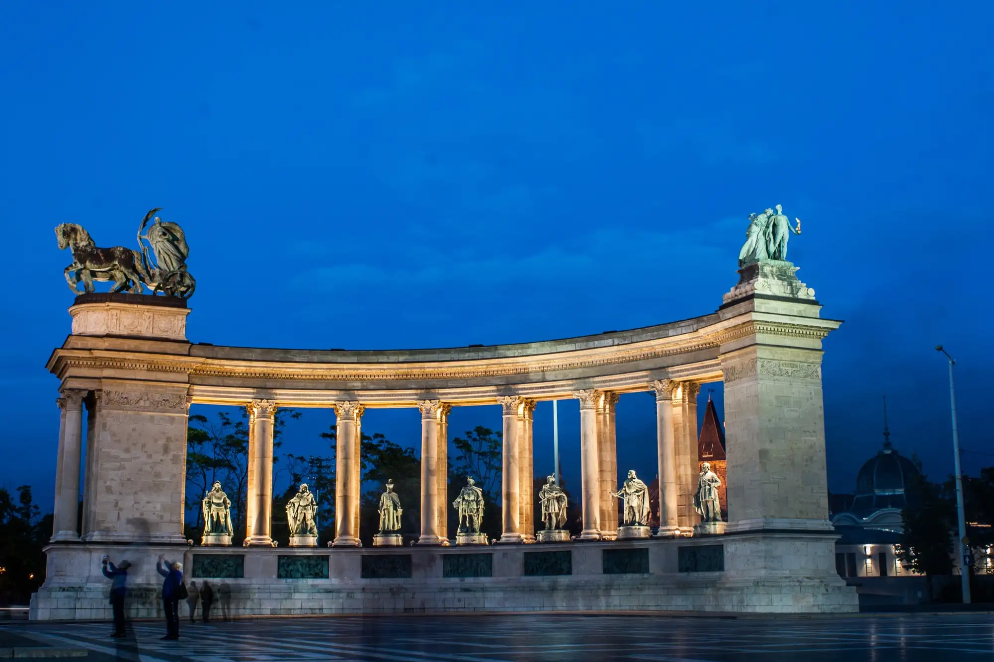 The right-hand colonnade of the Millennium Monument lit at dusk, seven bronze statues standing between its columns and the Peace chariot on its left-hand end