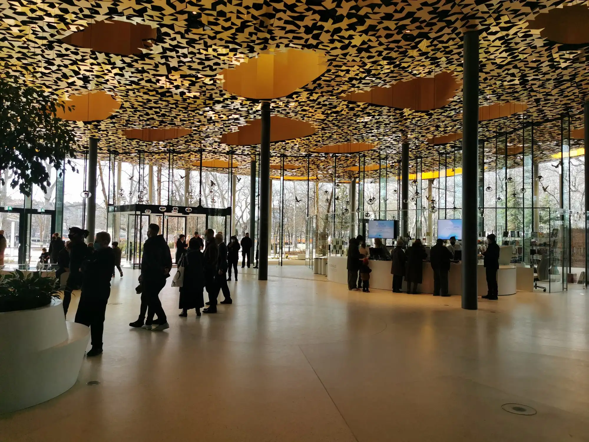 The undulating roof of the House of Hungarian Music from below, pierced by round openings and hung with golden leaf-patterned panels, with glass walls and visitors on the ground floor