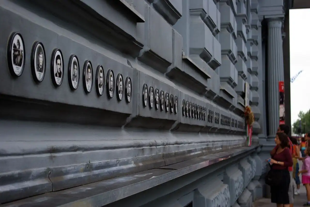 The row of oval portraits set into the base of the black frame of the House of Terror Museum, the museum's Wall of Heroes, with flowers left beneath