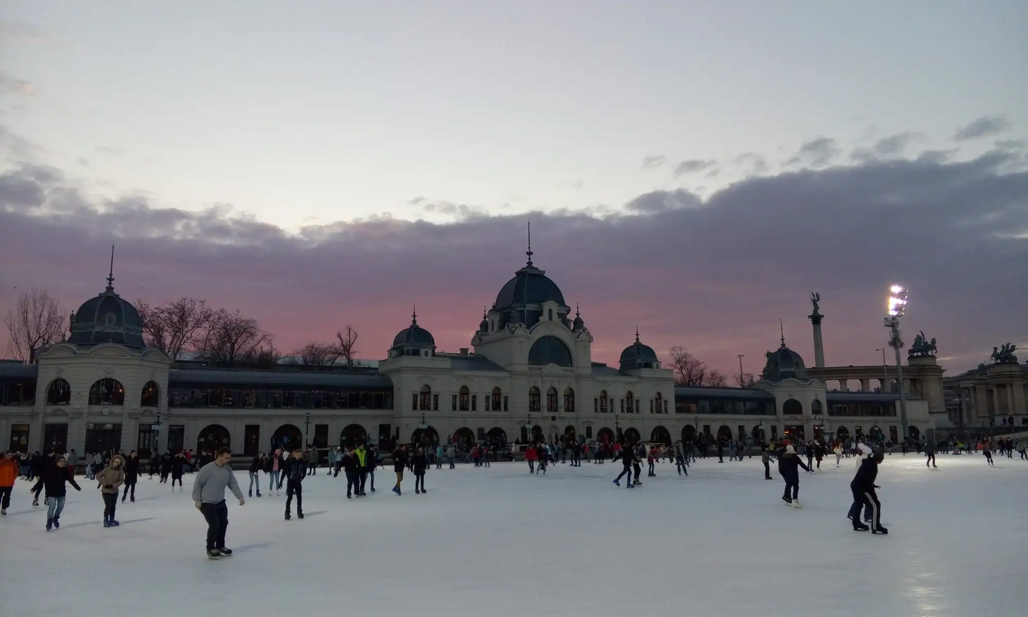 Skaters on the City Park Ice Rink at dusk, with the domed rink building lit behind them