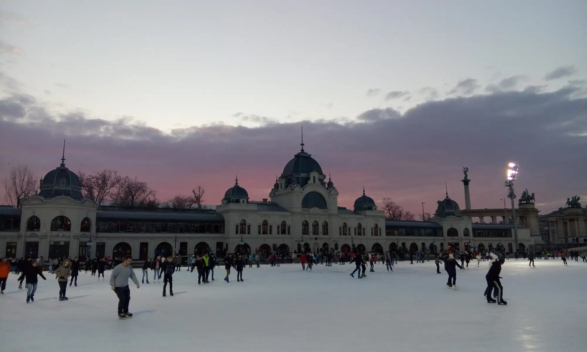Skaters on the City Park Ice Rink at dusk, with the domed Neo-Baroque rink building lit behind them