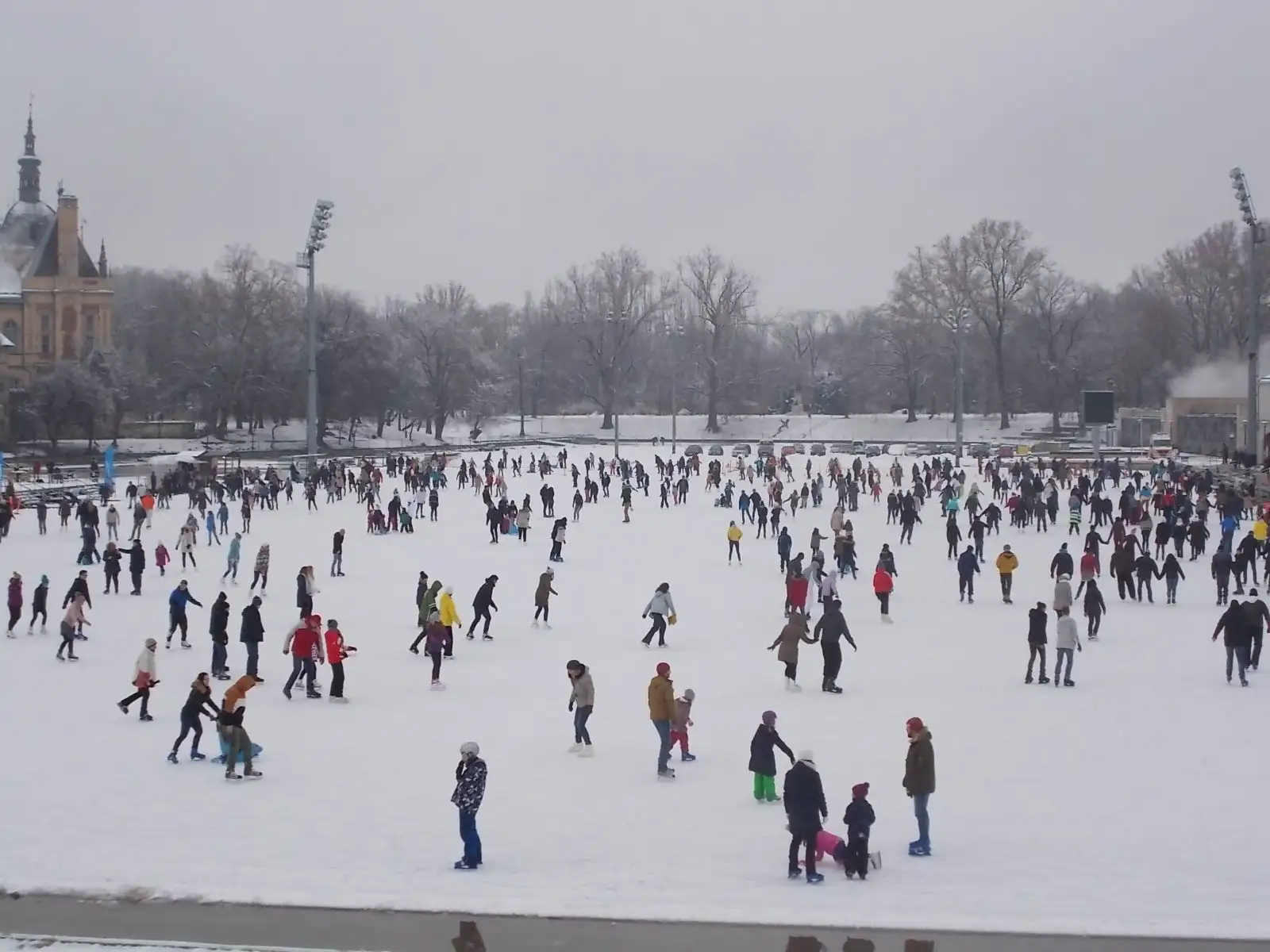 The City Park Ice Rink crowded with skaters on an overcast winter day, seen from the bridge, with bare trees around the lake