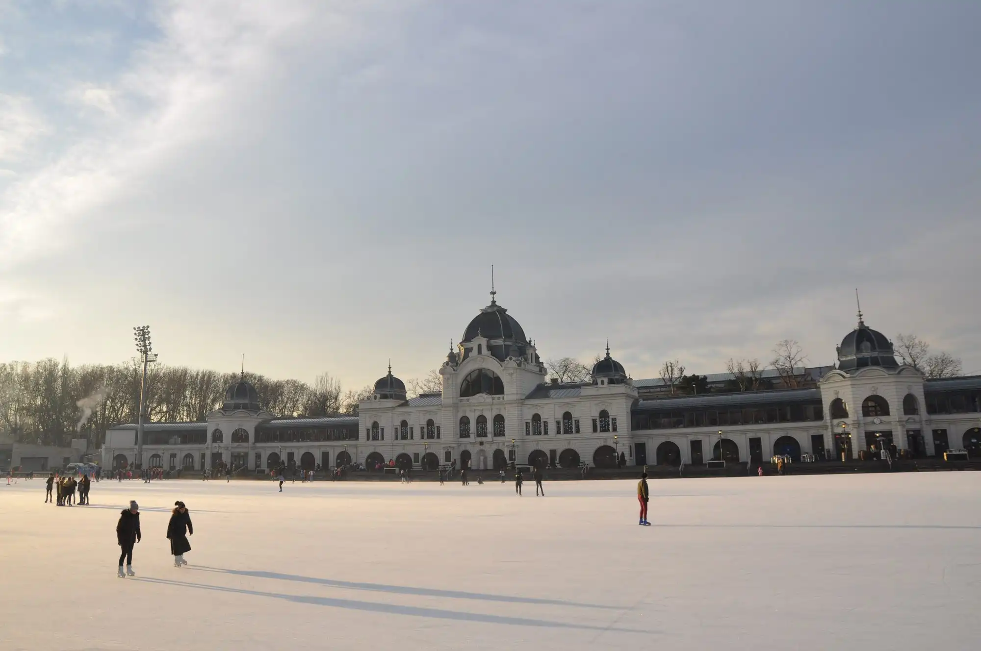 A few skaters spread across the wide, almost empty ice of the City Park Ice Rink on a clear winter day, with the domed rink building on the far side