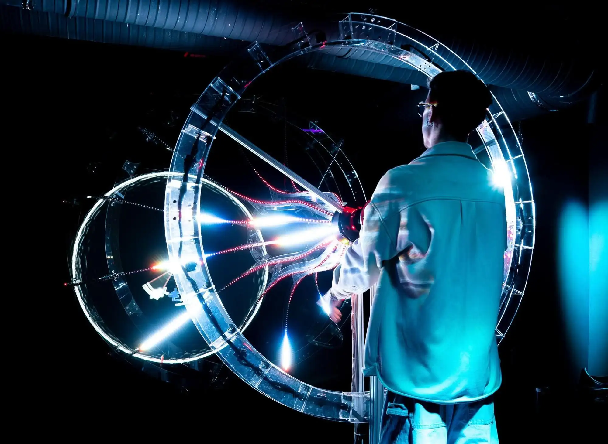 A visitor operating a large circular light sculpture at Cinema Mystica in Budapest, its rings lit with white and red LEDs in a dark room