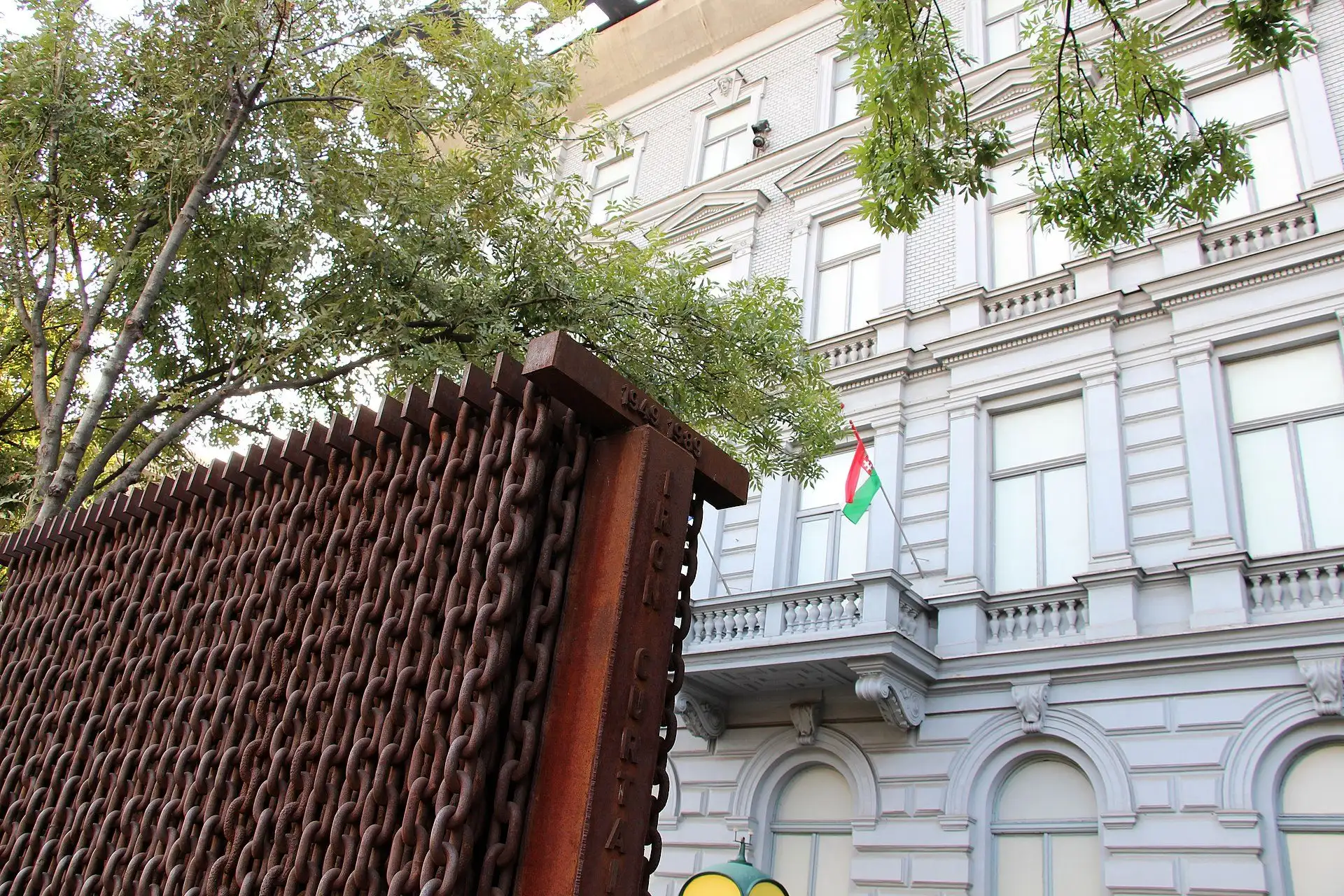 The Iron Curtain sculpture on the pavement of Andrássy út, a wall of rusted chains hung from steel girders, with the facade of the House of Terror Museum behind