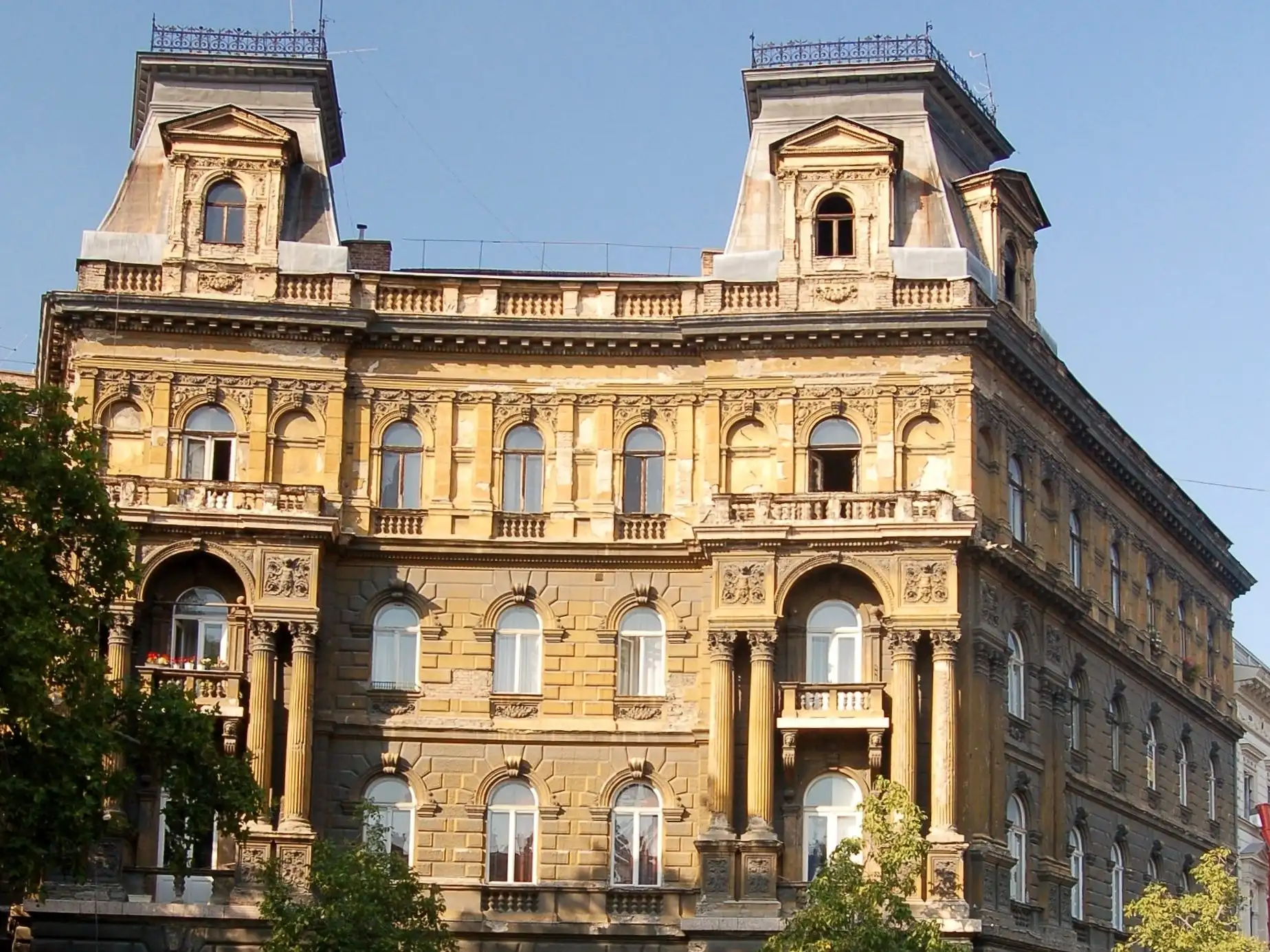 The Hübner-udvar at Kodály körönd in Budapest, a yellow Neo-Renaissance block with corner pavilions, seen across the planted circus in low sun