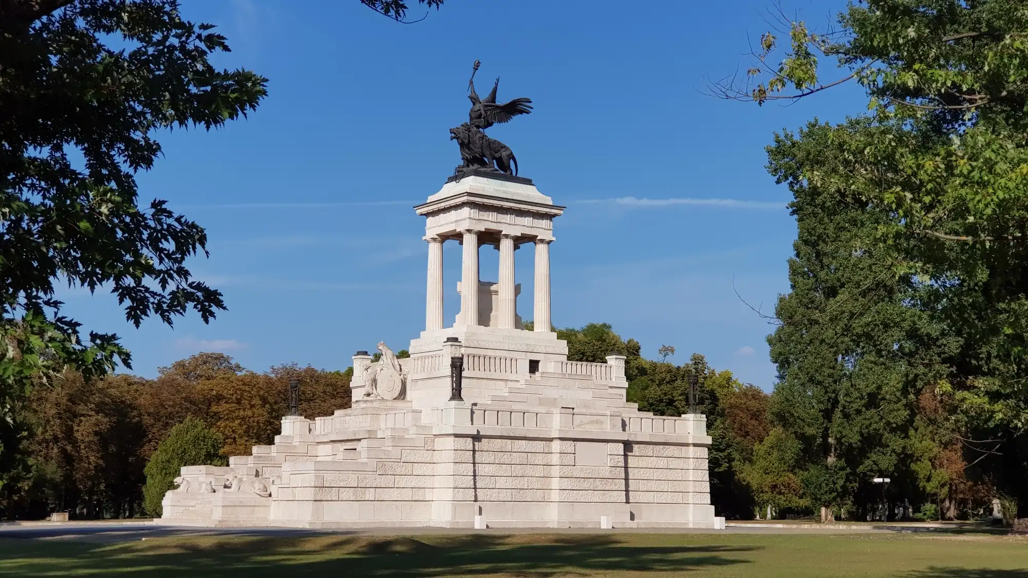 The white stone Kossuth Mausoleum in Fiumei Road Cemetery, a stepped base and colonnade topped by a bronze winged figure, framed by trees under a blue sky