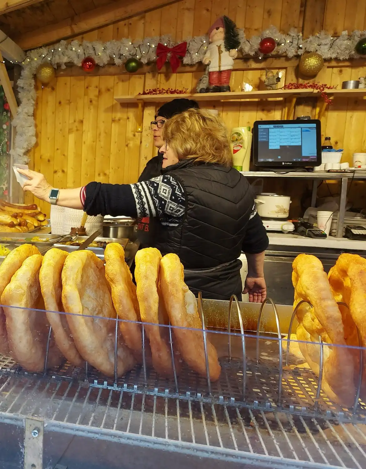 Freshly fried lángos stacked upright in a rack at a wooden food stall at the Advent Bazilika market, with a cook handing over an order