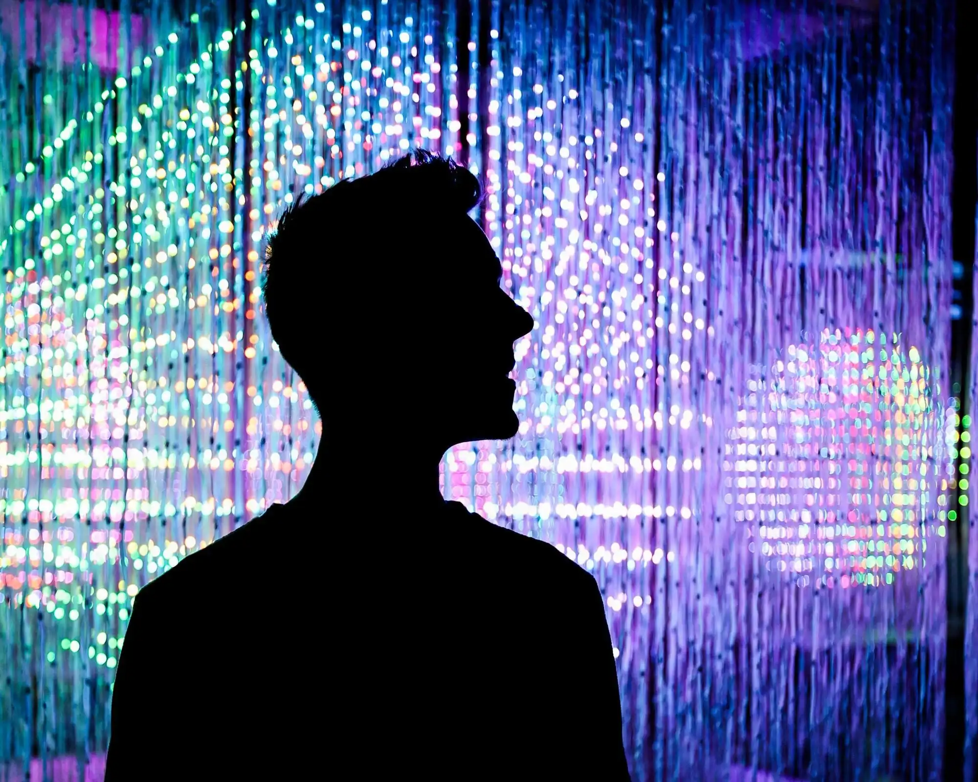 A visitor silhouetted against a curtain of hanging light strands glowing blue, pink and green in a dark room at Cinema Mystica in Budapest