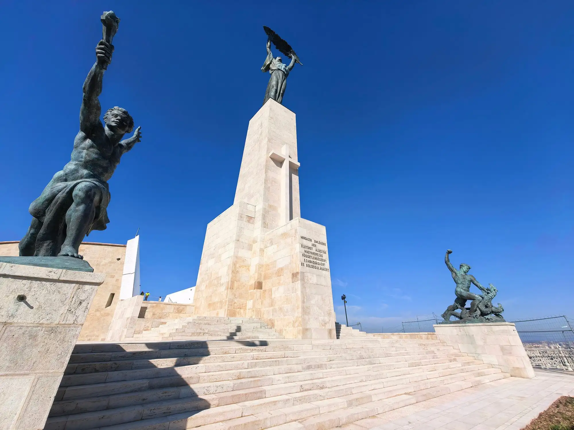The Liberty Statue on Gellért Hill, the bronze Genius holding a palm leaf on its tall stone pedestal, with a cross set into the pedestal and the two bronze side figures on their plinths