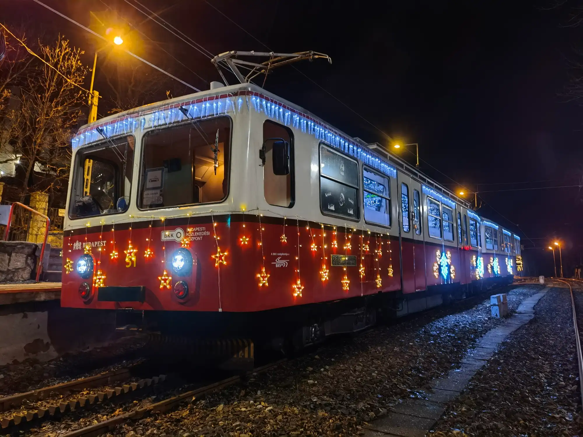 The red and white cogwheel train in the Buda hills at night, hung with blue icicle lights and gold stars, with snow beside the track
