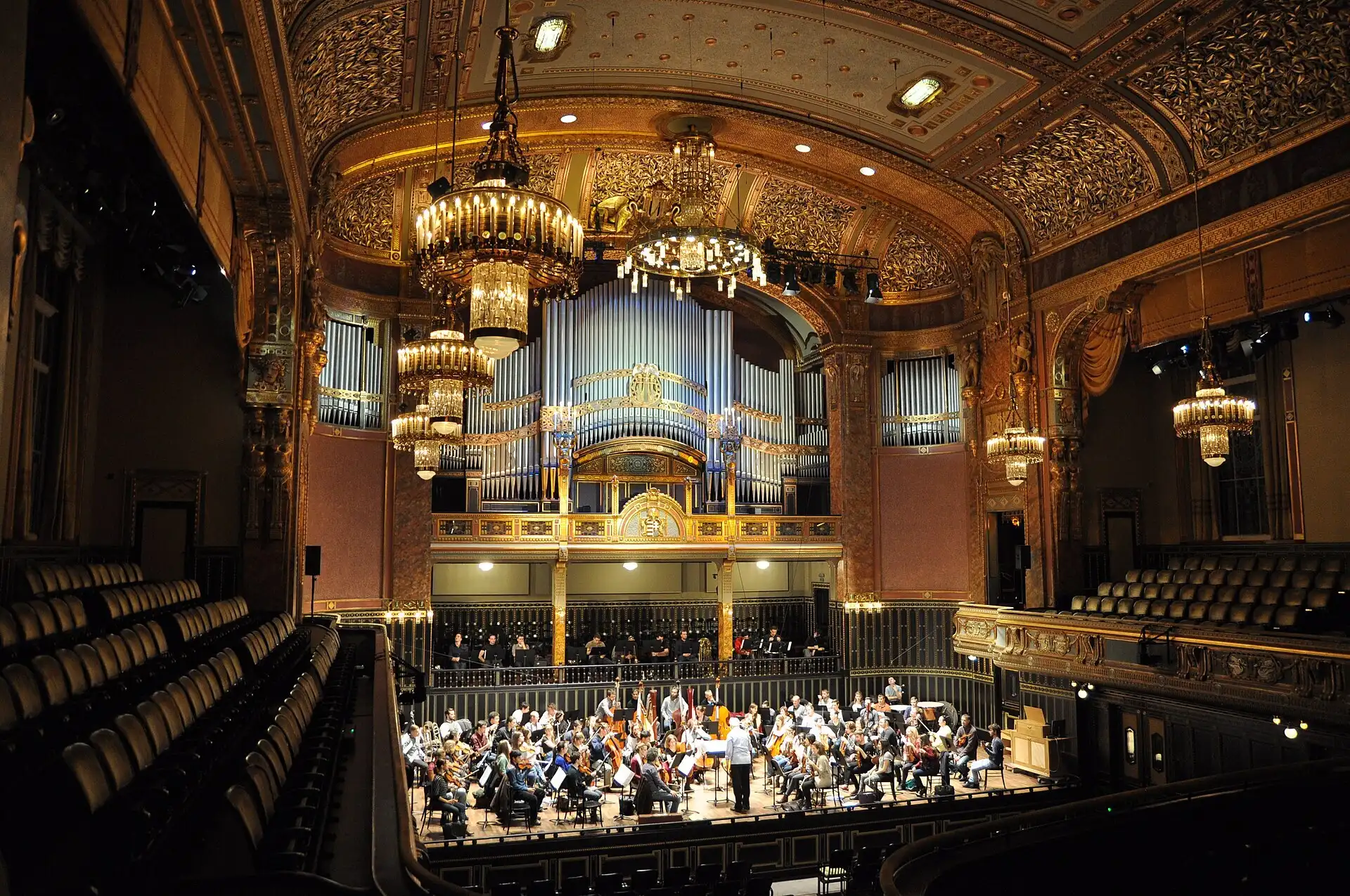 The Grand Hall of the Liszt Academy in Budapest, its gilded Art Nouveau tiers and chandeliers above an orchestra rehearsing in front of the organ