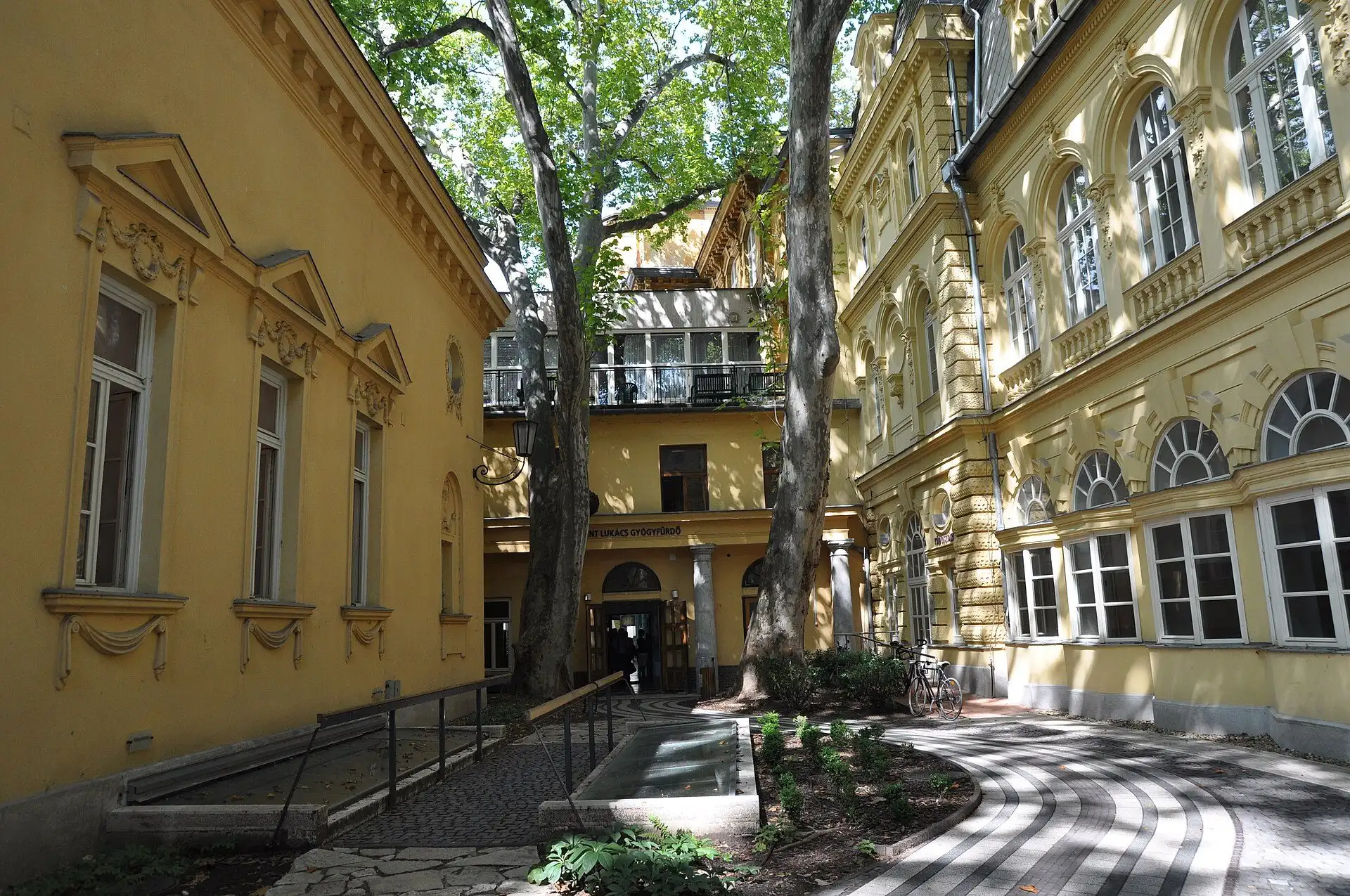 The courtyard at Lukács Thermal Bath, with plane trees between the yellow wings and the entrance door under a sign at the back