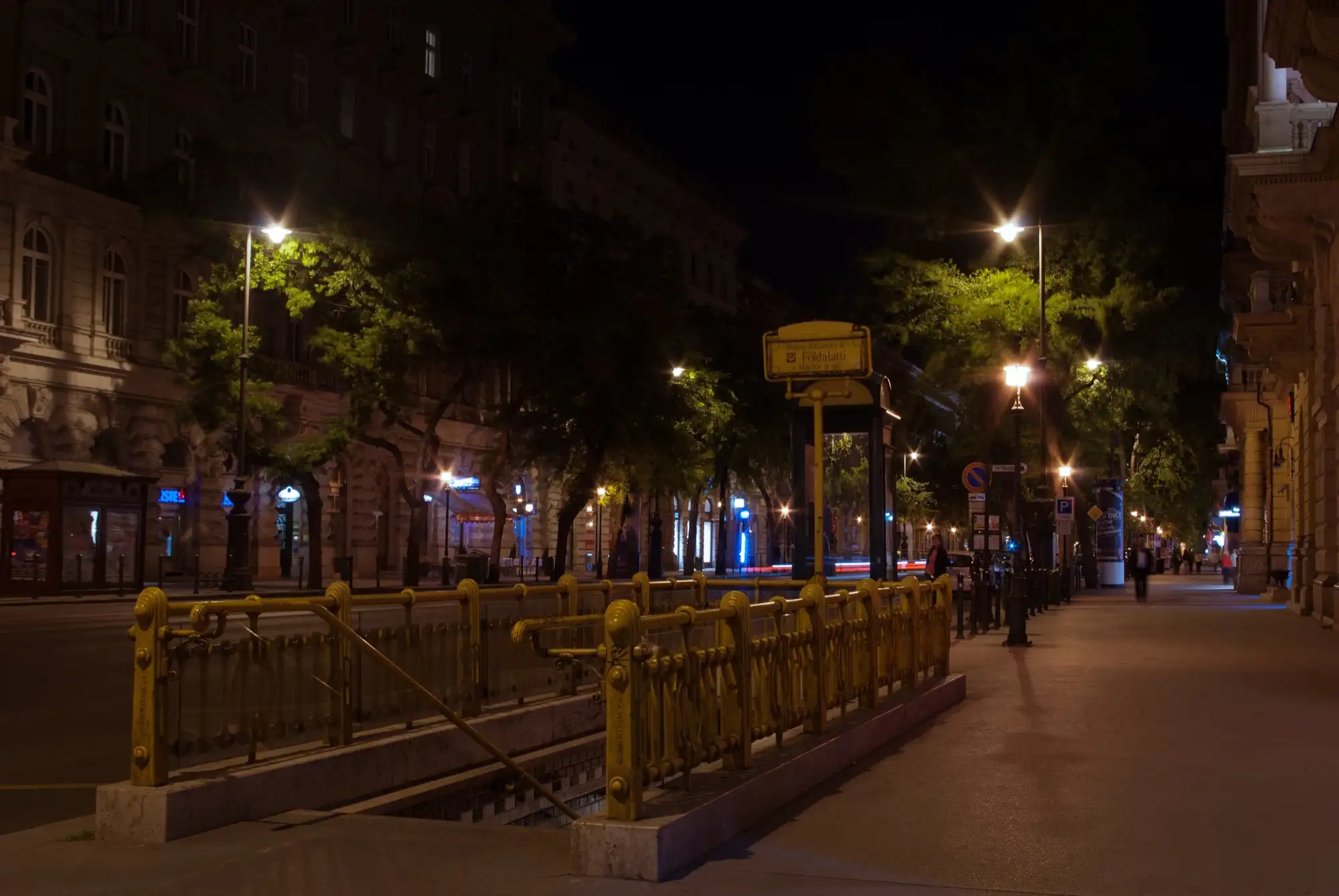 The yellow-painted iron railings round the M1 stairway at Bajcsy-Zsilinszky út on Andrássy Avenue at night, with the station sign on a post