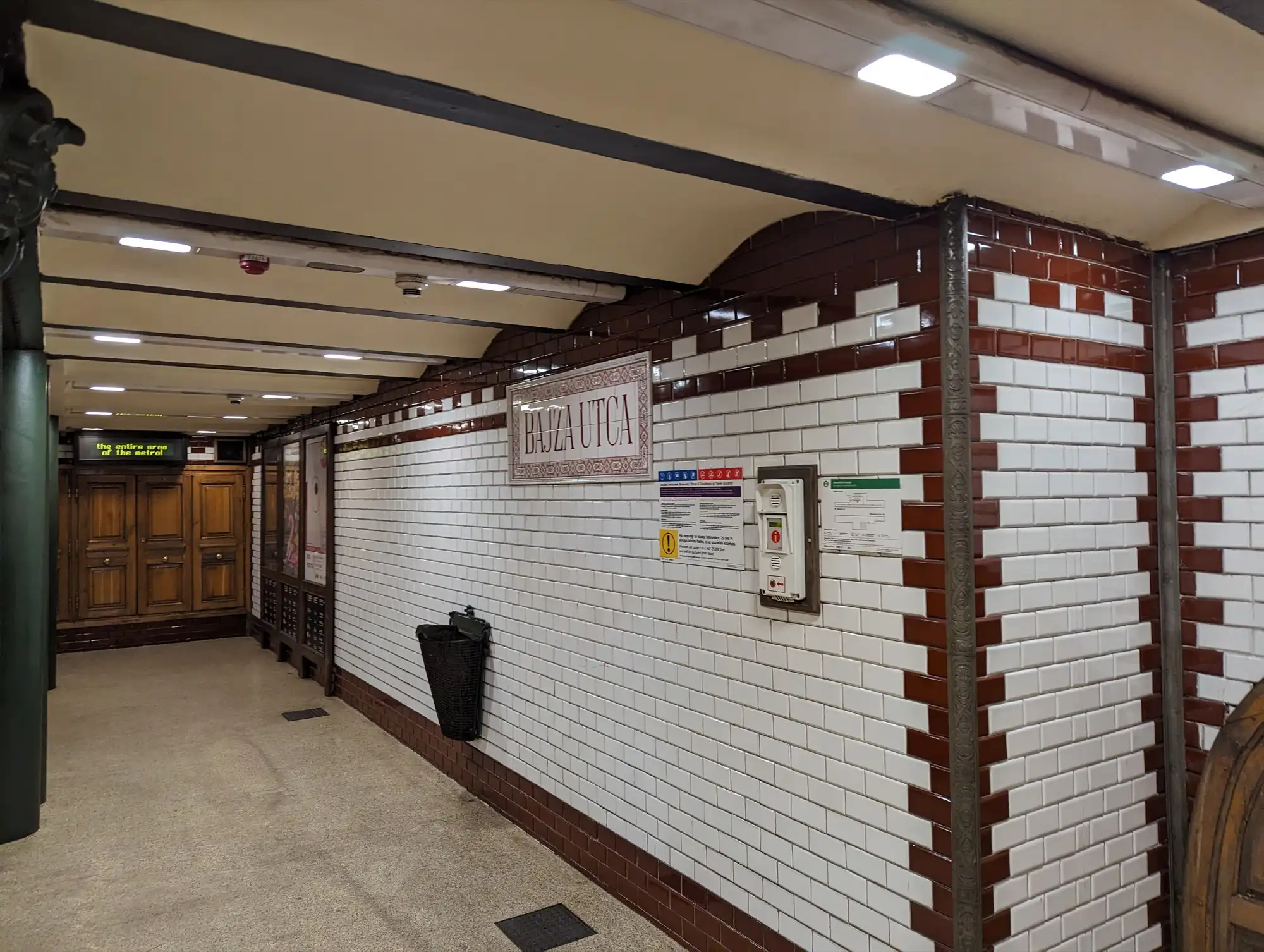 The tiled wall at Bajza utca station on the M1, white tiles framed in dark red with the station name on a tiled panel and timber doors at the end
