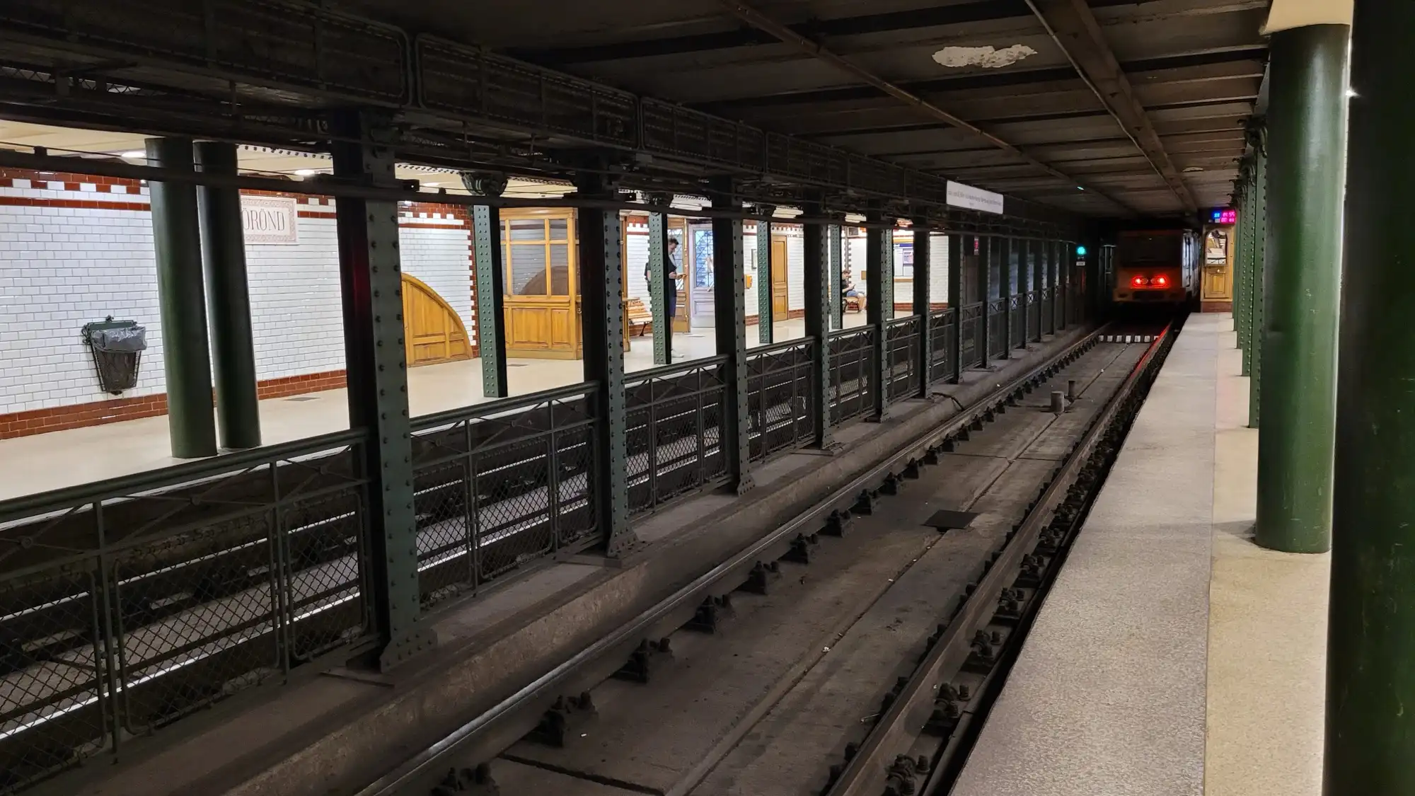 The platform at Kodály körönd station on the M1, a row of green riveted columns along the track and white tiled walls trimmed in dark red