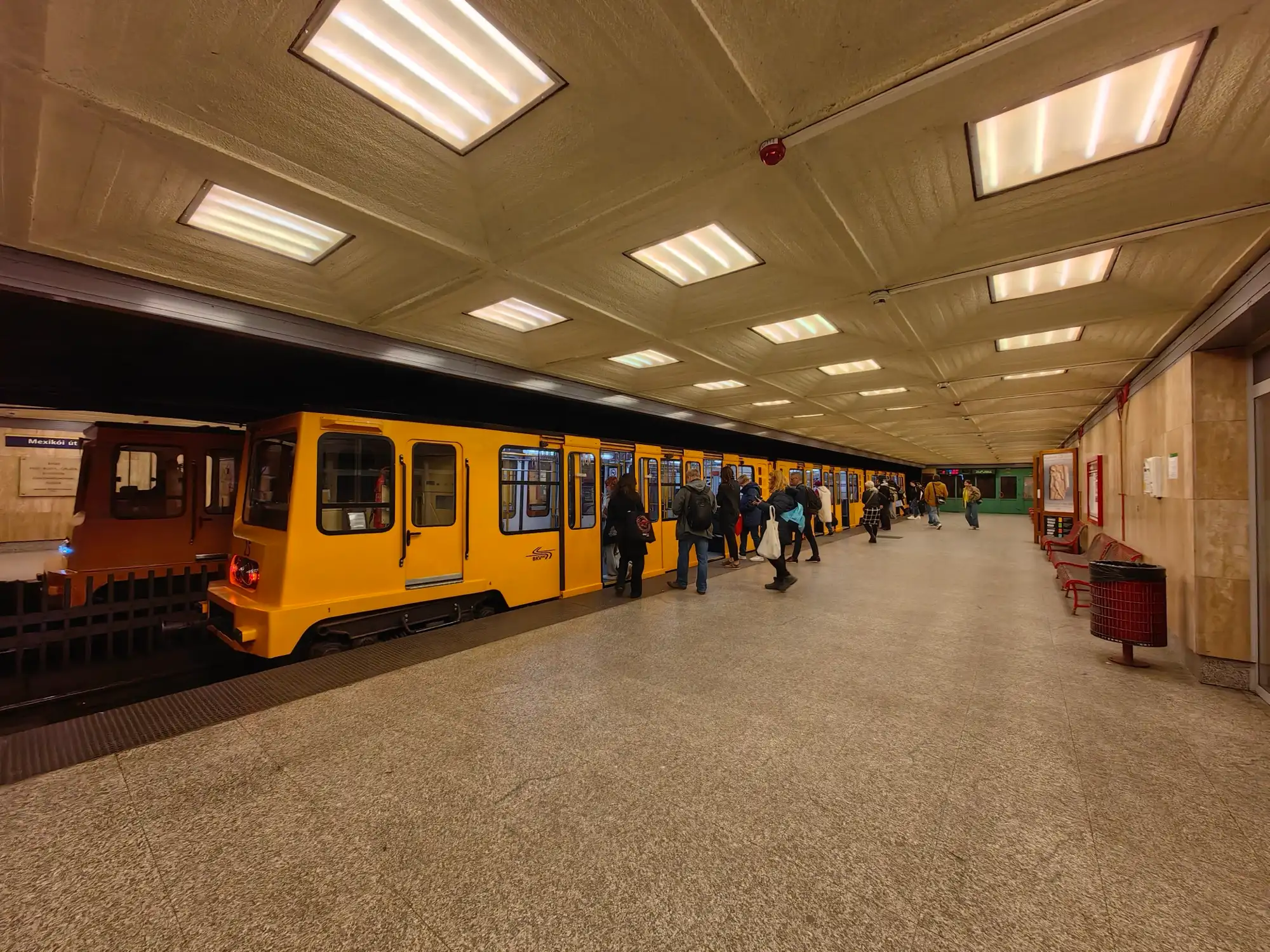 Yellow M1 trains at the wide platform of the Mexikói út terminus in Budapest, under a panelled ceiling with strip lights