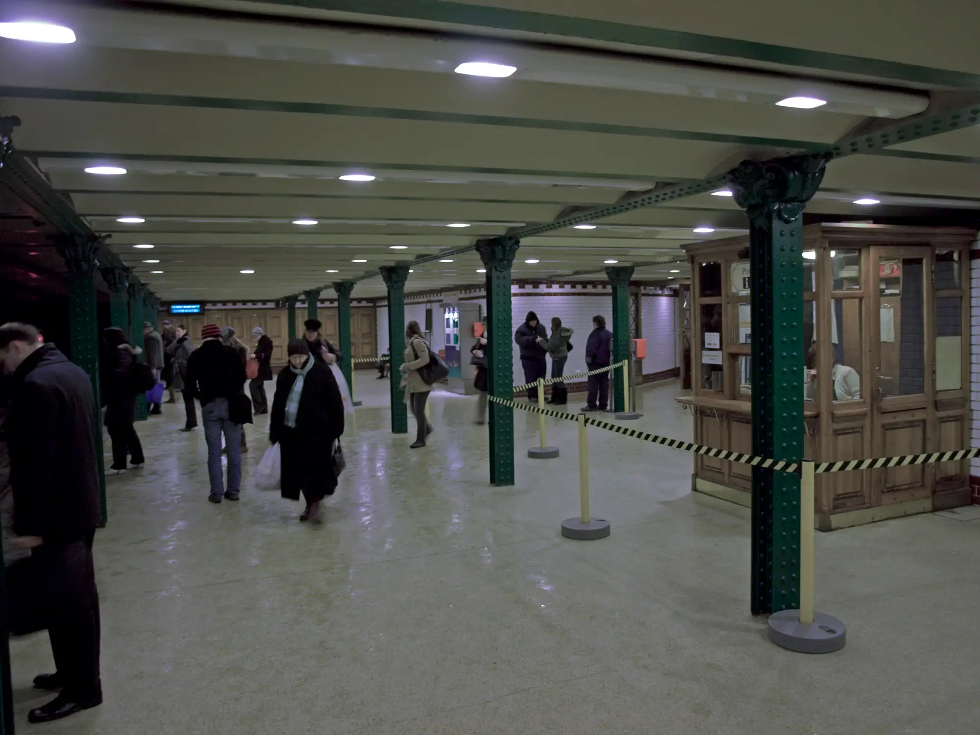 Inside Oktogon station on the M1, green riveted iron columns down the middle and a timber booth on the right, with passengers walking through