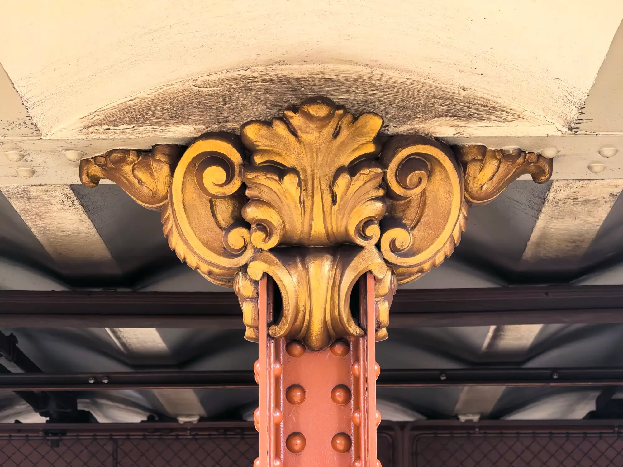 A gilded capital at the top of a brown-painted riveted iron column under the ceiling of Opera station on the M1 in Budapest