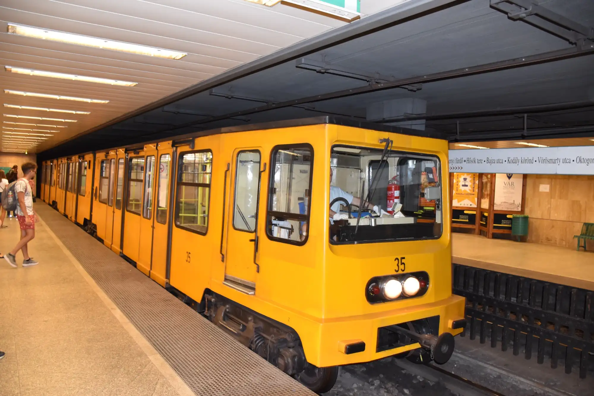 A yellow M1 train at the platform of Széchenyi fürdő station in City Park, with plain panelled walls and a flat ceiling