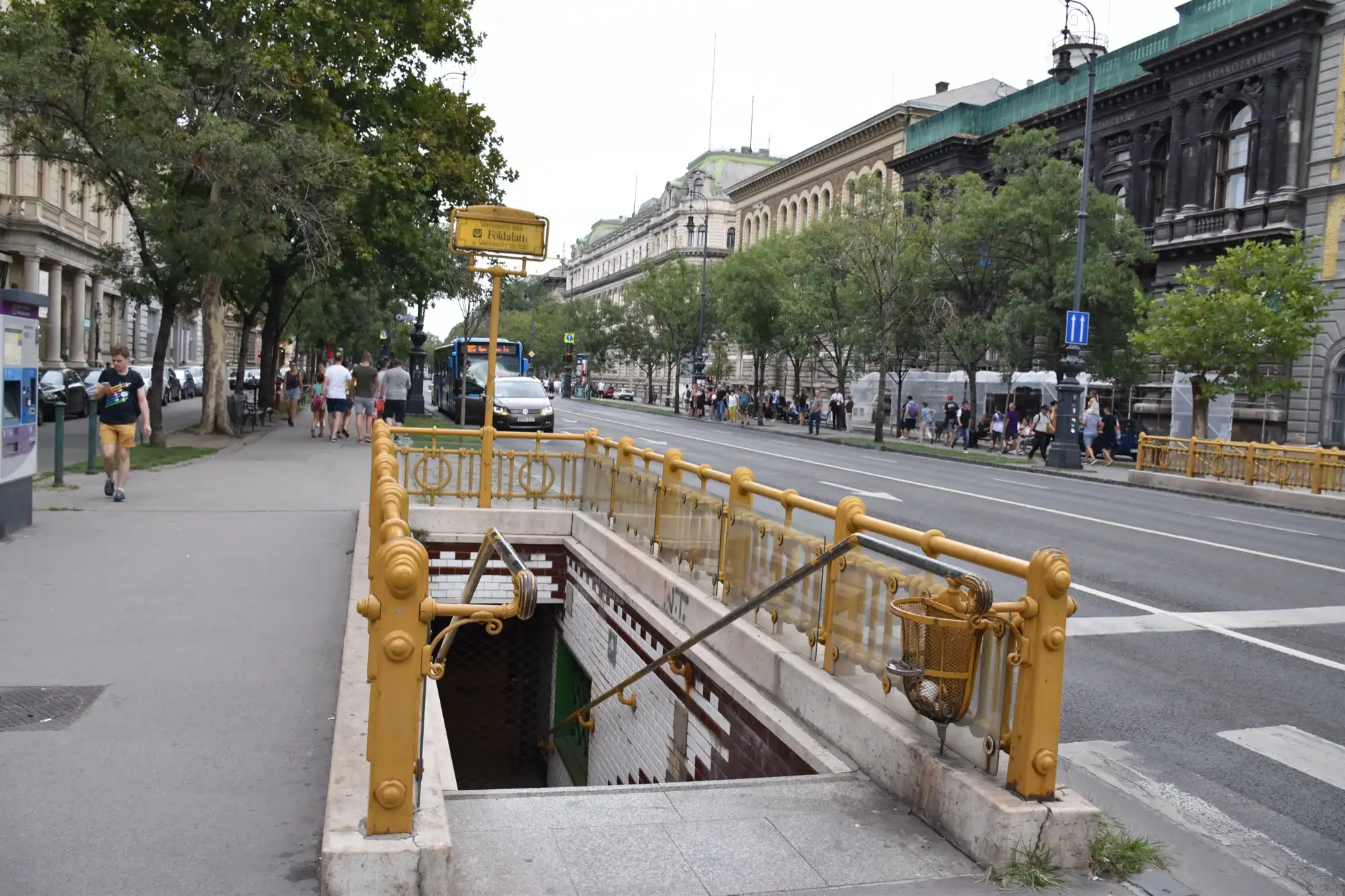The stairway down to Vörösmarty utca station on Andrássy Avenue, with yellow-painted iron railings and the station sign on a post, the avenue's trees and facades behind