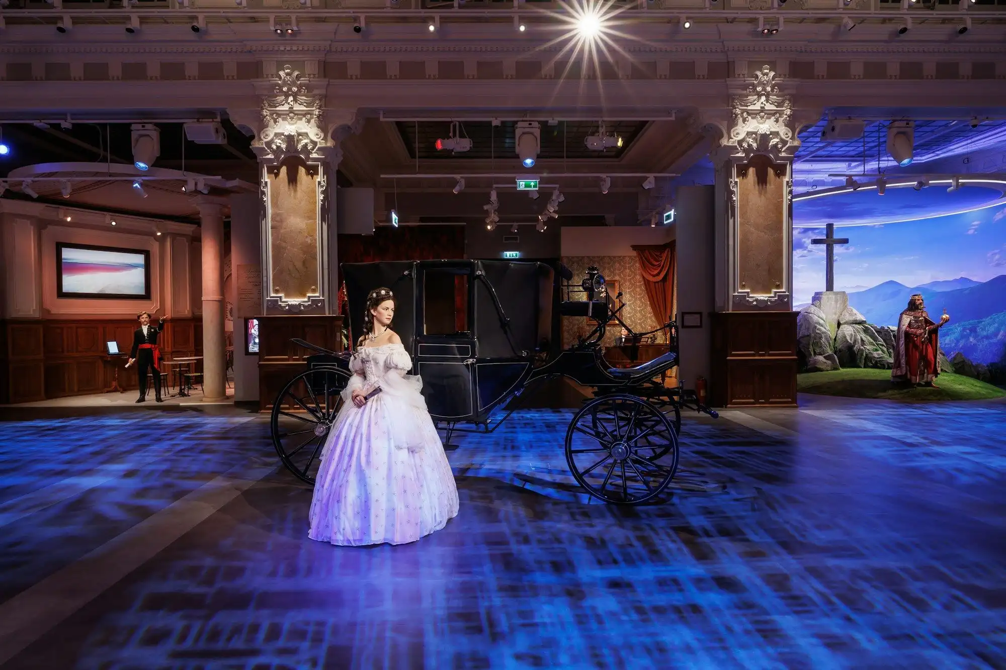 The Sissi wax figure in a white gown beside her carriage in the Hungarian Spirit zone of Madame Tussauds Budapest, with the King Stephen set behind