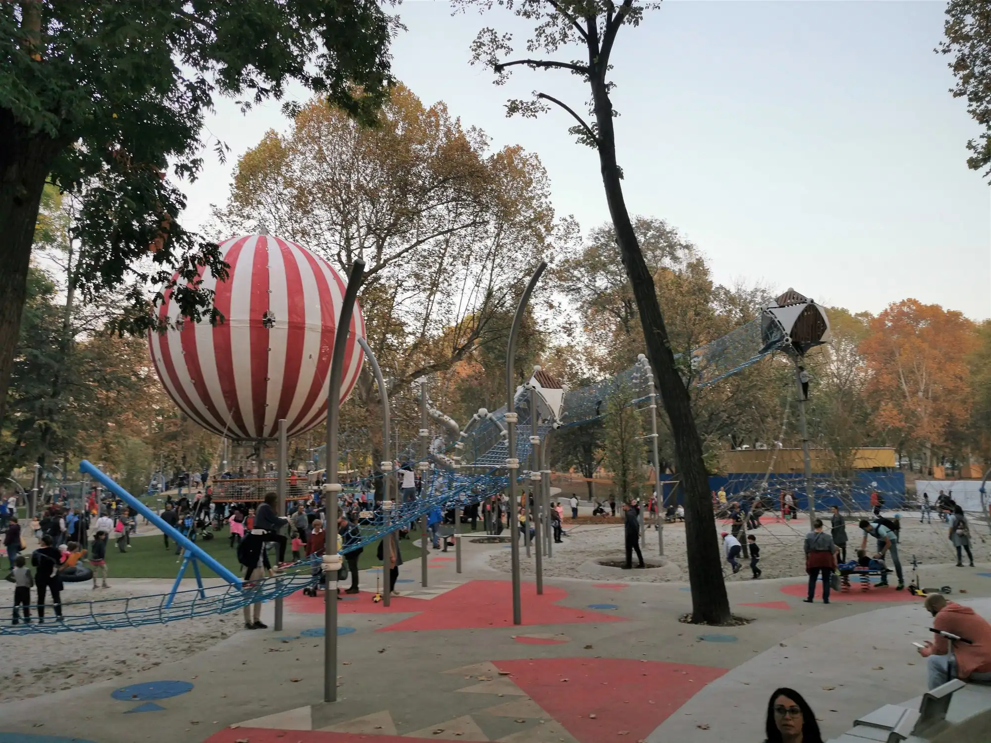 The Main Playground in Budapest's City Park, with children on climbing equipment and a tall red and white striped balloon-shaped climbing frame among autumn trees