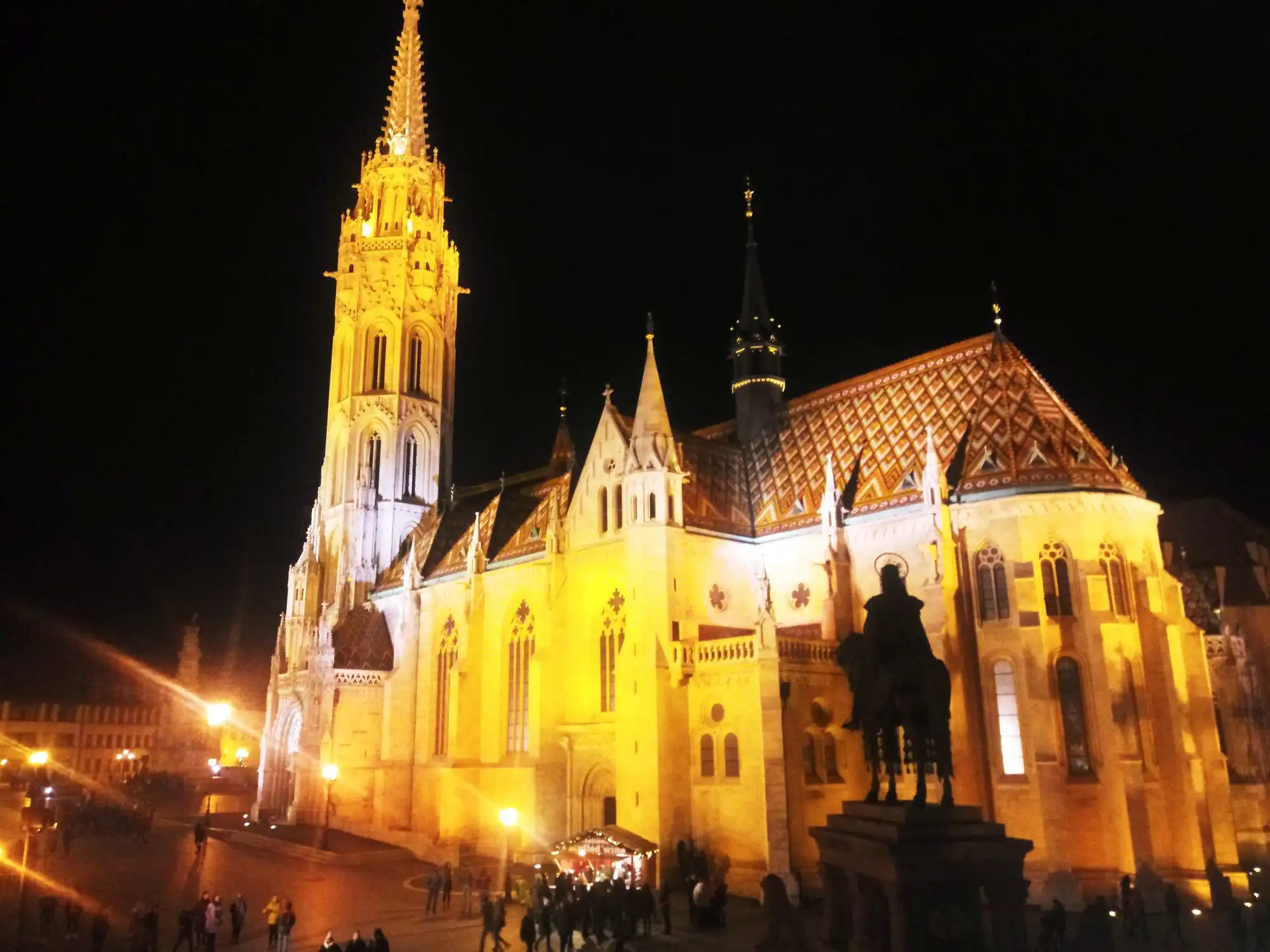 Matthias Church floodlit at night from Szentháromság tér, with the St Stephen statue in silhouette and the tiled roof lit up