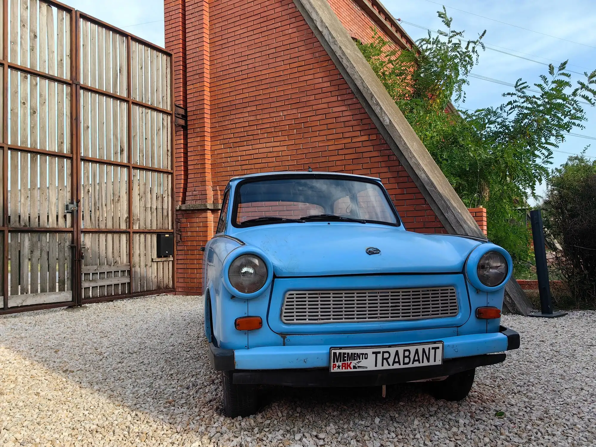 A pale blue Trabant parked on gravel beside the brick wall of Memento Park